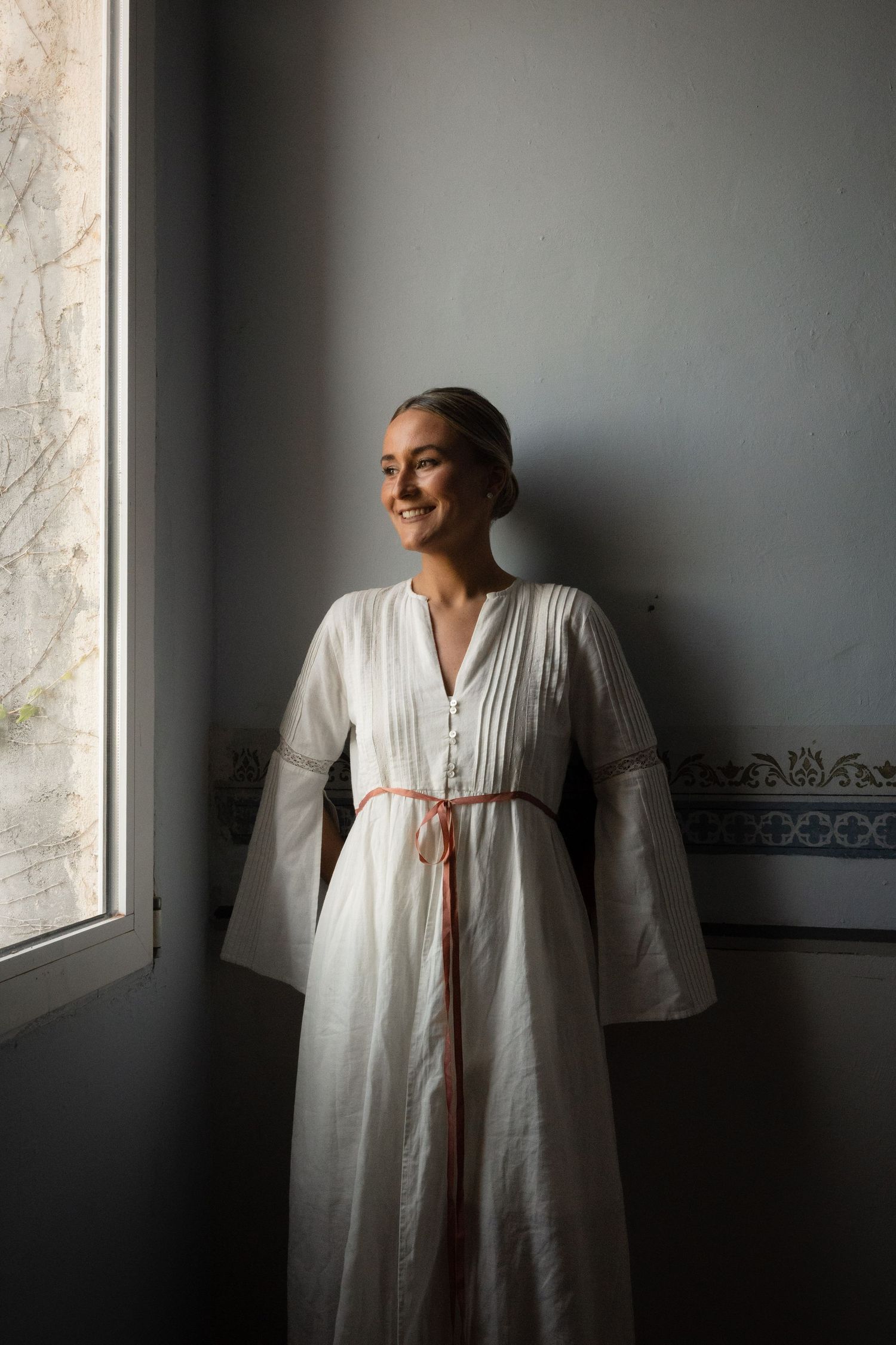 Figure in flowing white dress with pink sash stands by window in natural light.