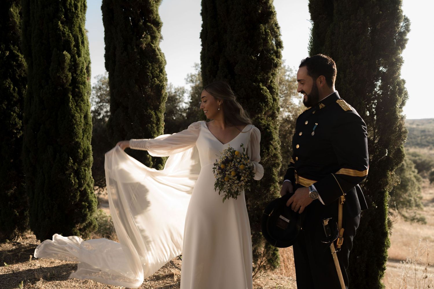 Flowing white bridal veil catches sunlight between tall cypress trees in an outdoor setting.