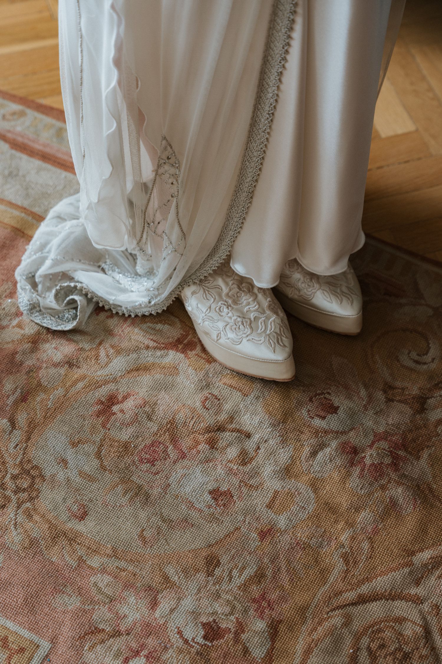 Close-up view of white wedding dress hem and delicate lace details against an ornate beige and pink patterned carpet.