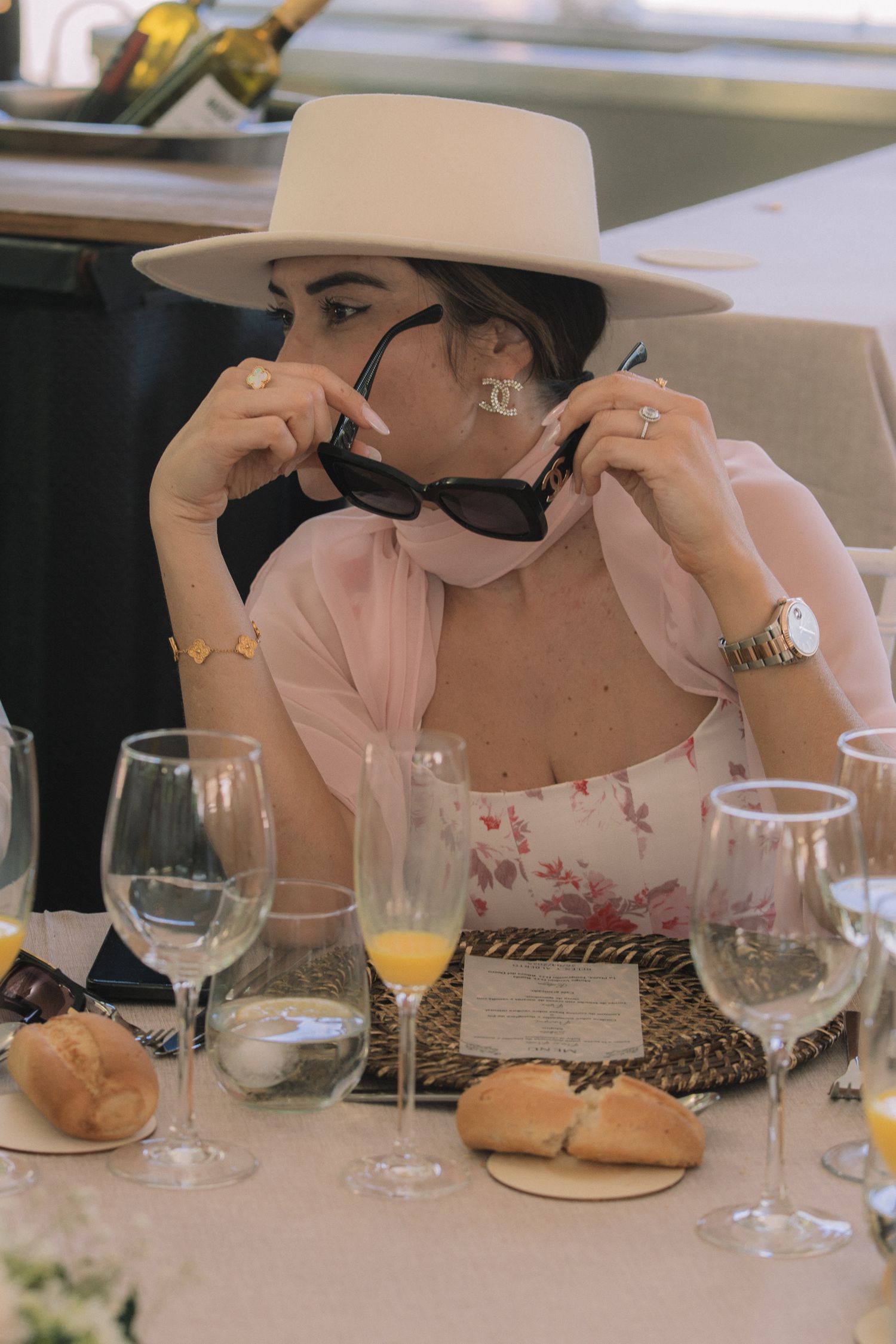 Elegant diners in wide-brimmed hats seated at a formal table with wine glasses and place settings.