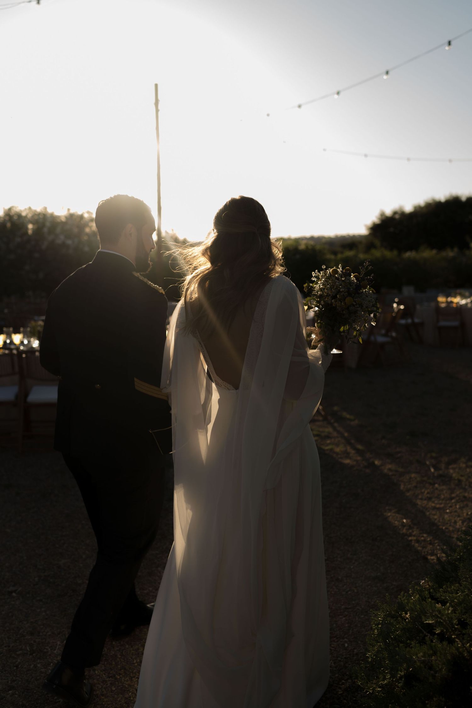 Backlit wedding couple walking together at sunset with string lights visible overhead.