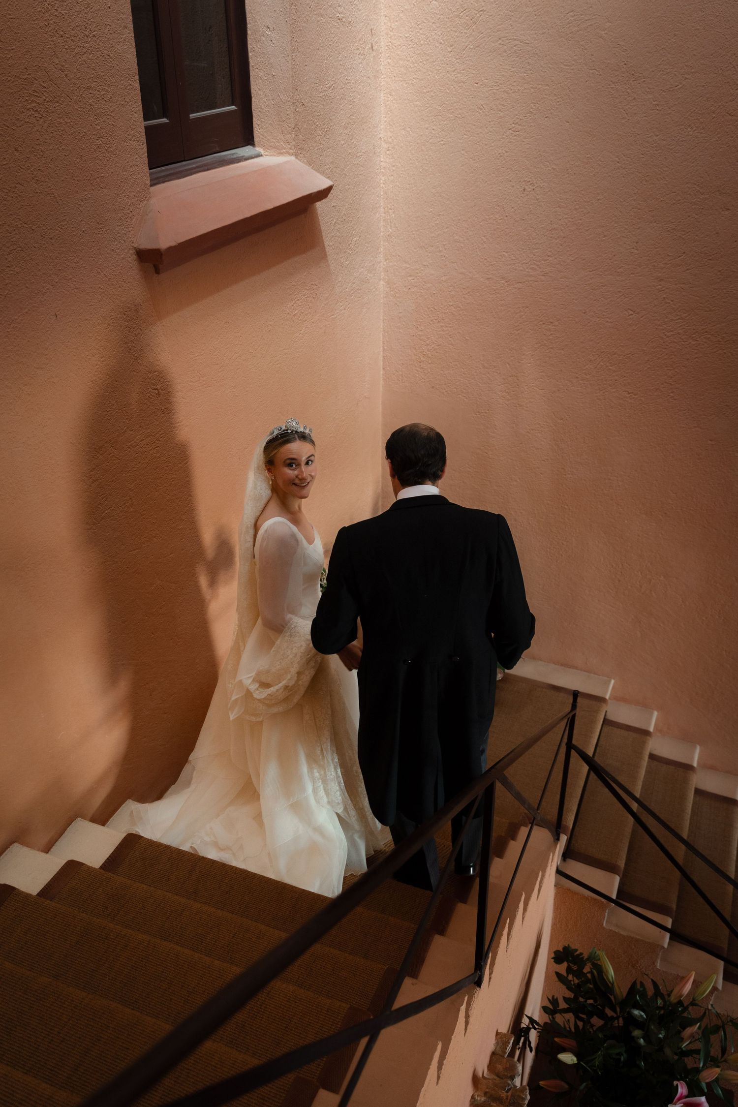 A couple descends a staircase in elegant wedding attire against a warm-toned wall.