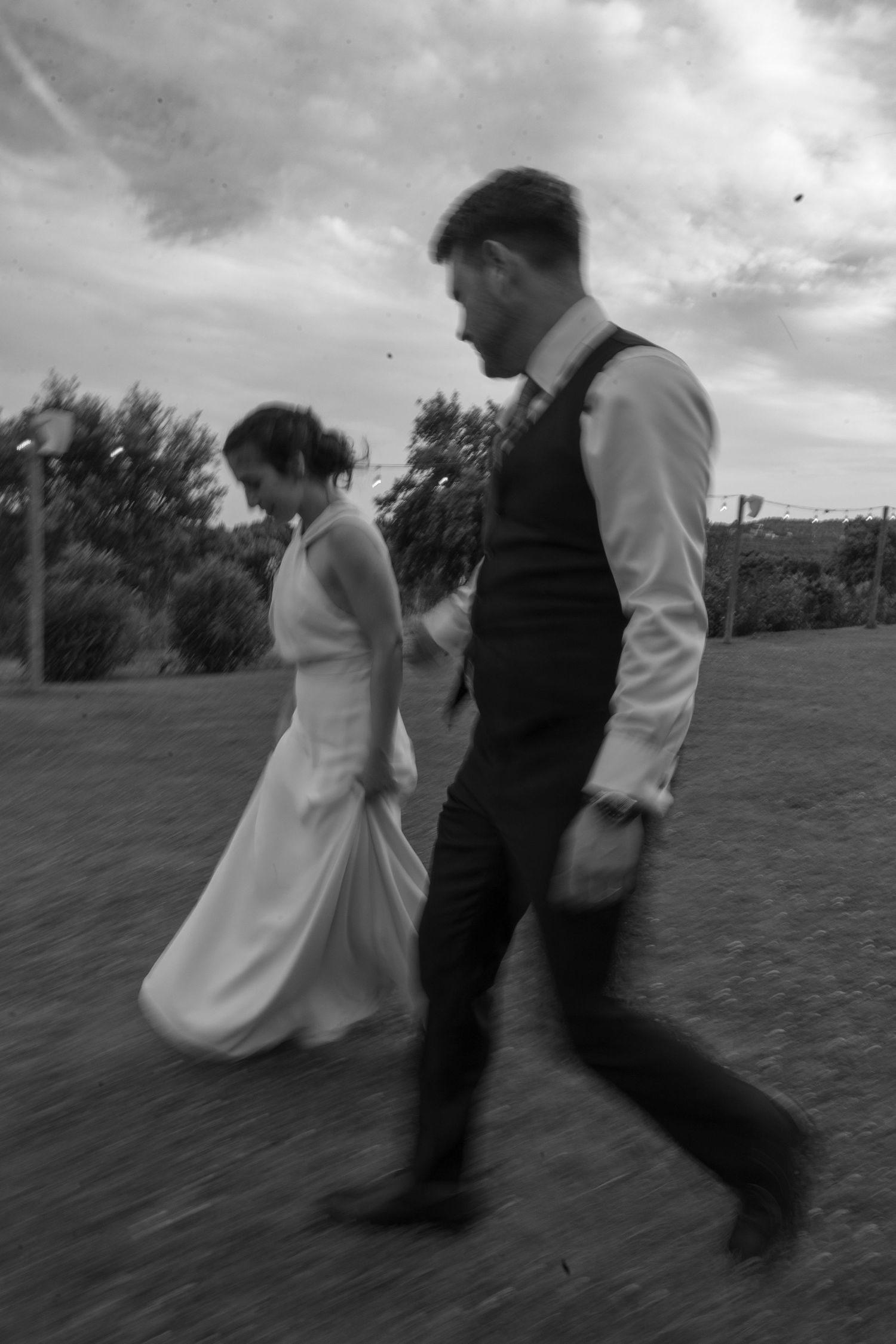 Wedding couple running and playing in a grassy field under cloudy skies.