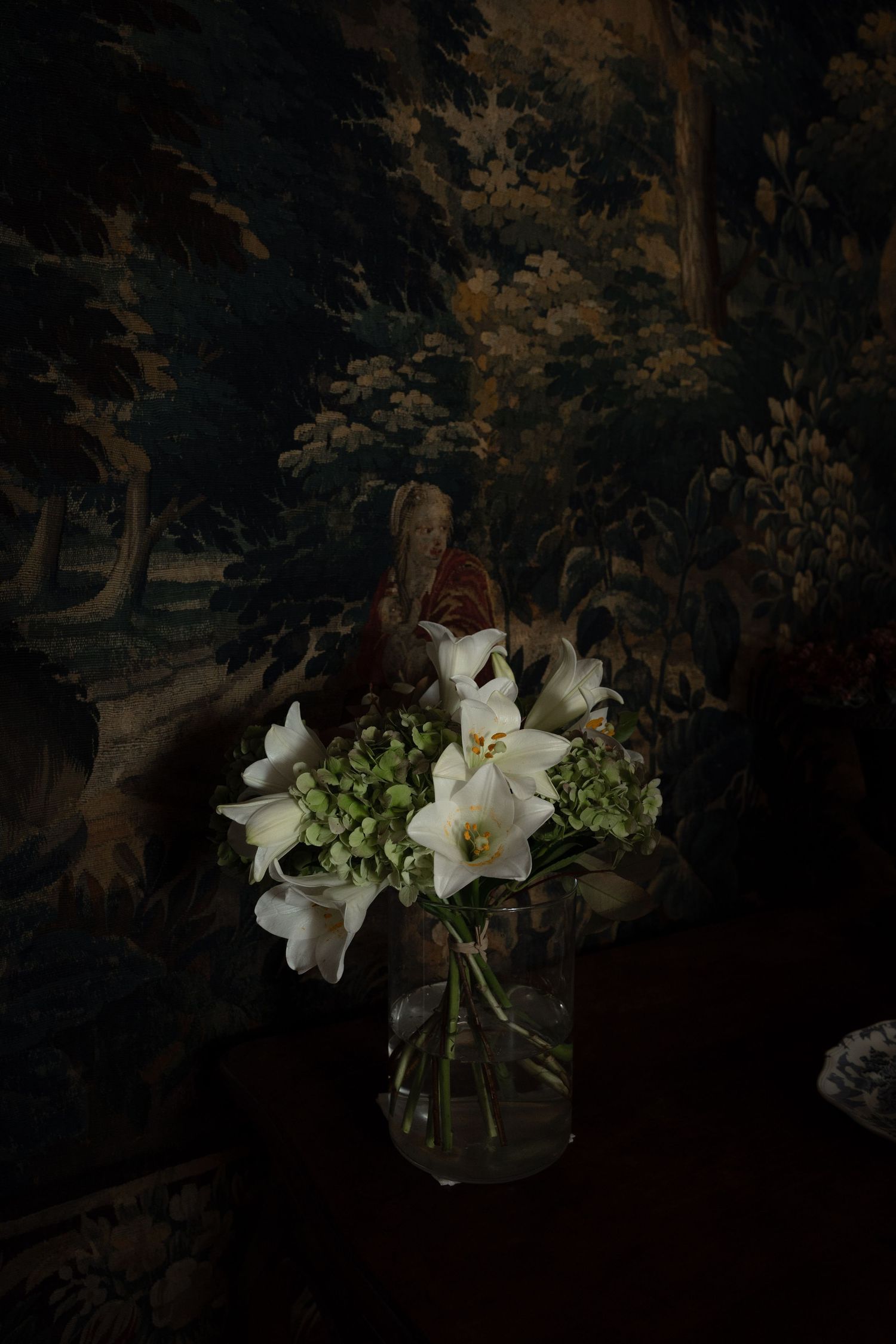 White lilies and hydrangeas in glass vase against dark floral tapestry background.