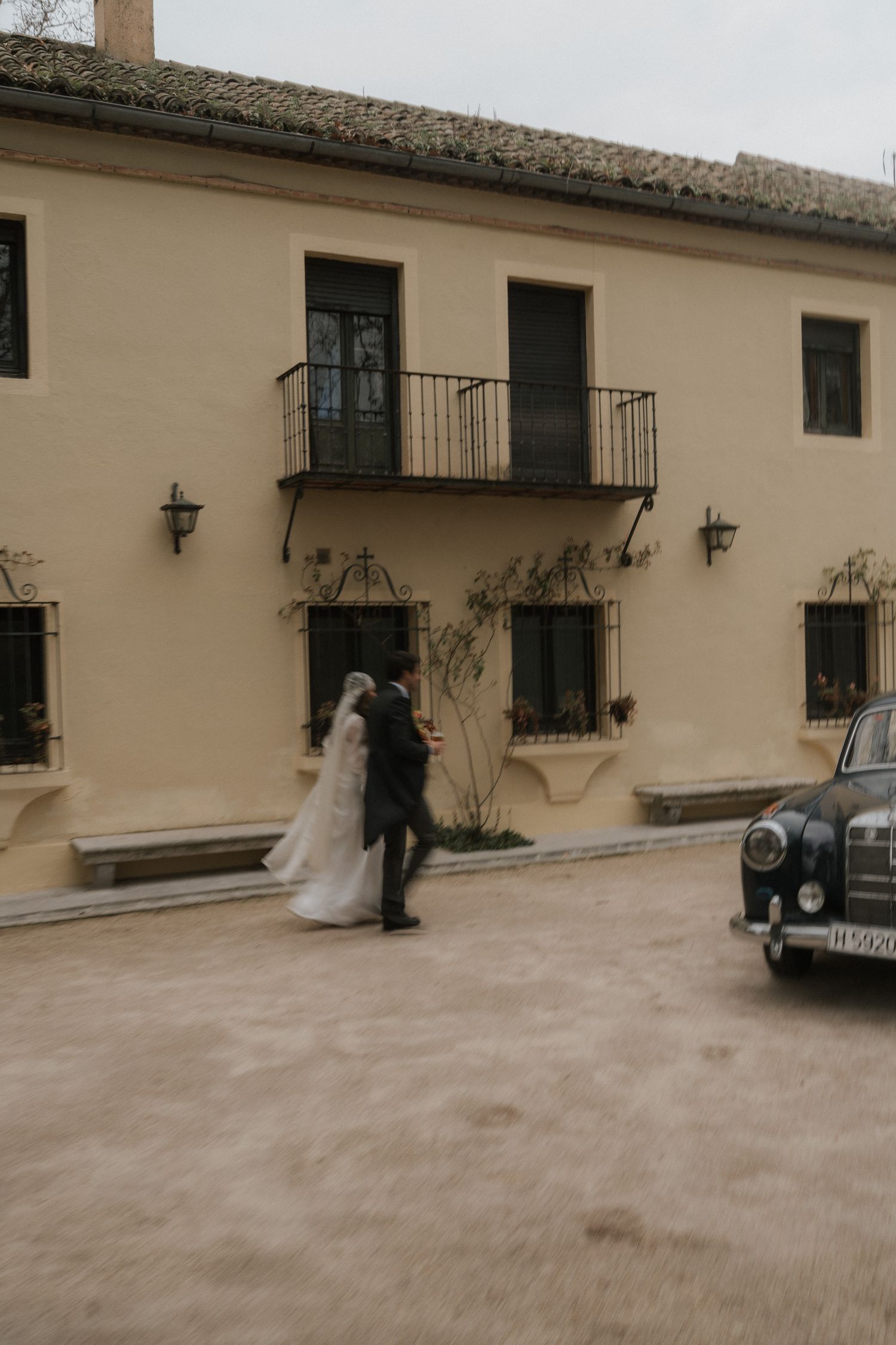 Wedding couple poses near a rustic building exterior with vintage car in the courtyard.