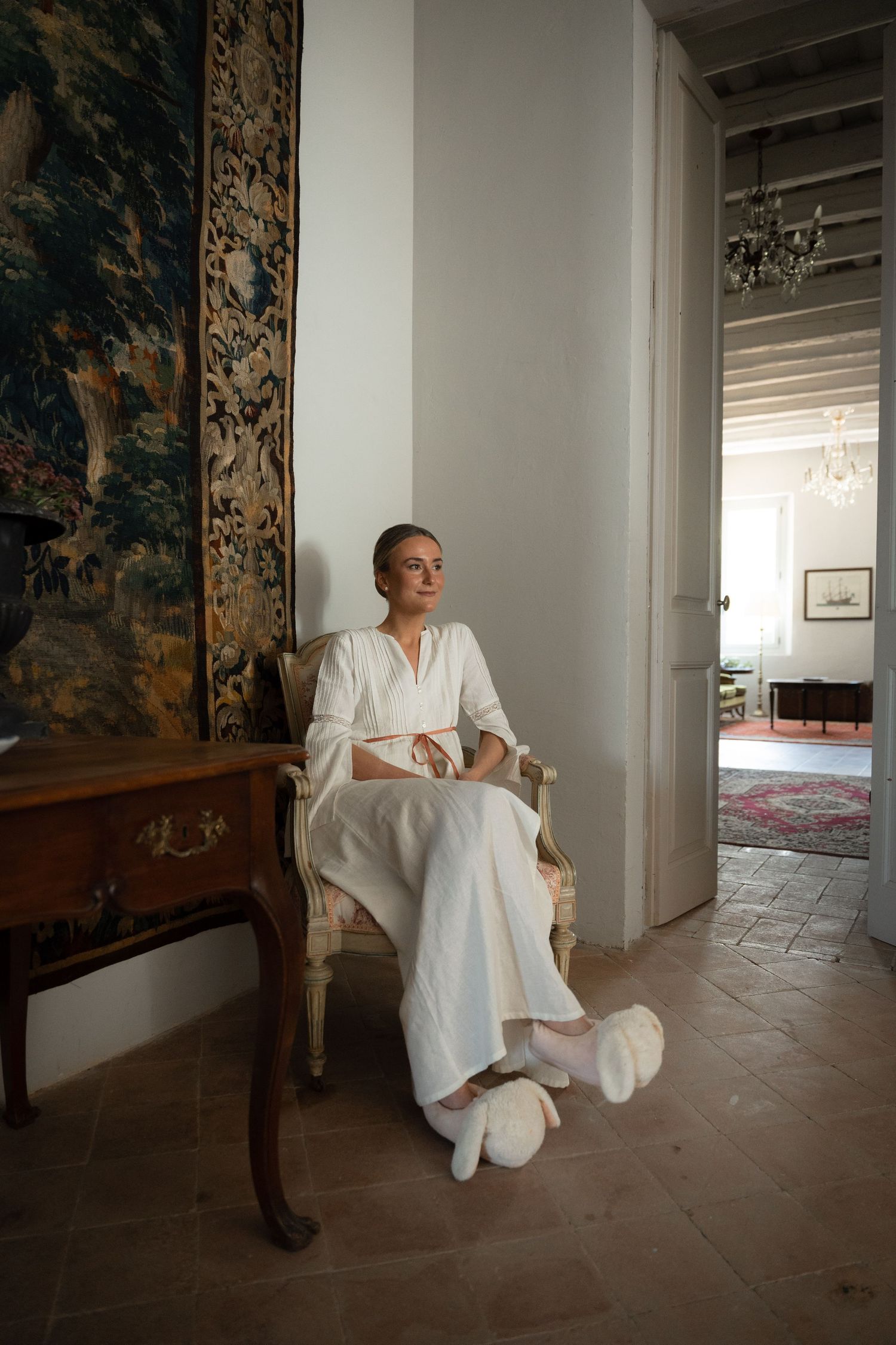 Elegant figure in white dress seated in vintage chair beside decorative tapestry in sunlit interior.