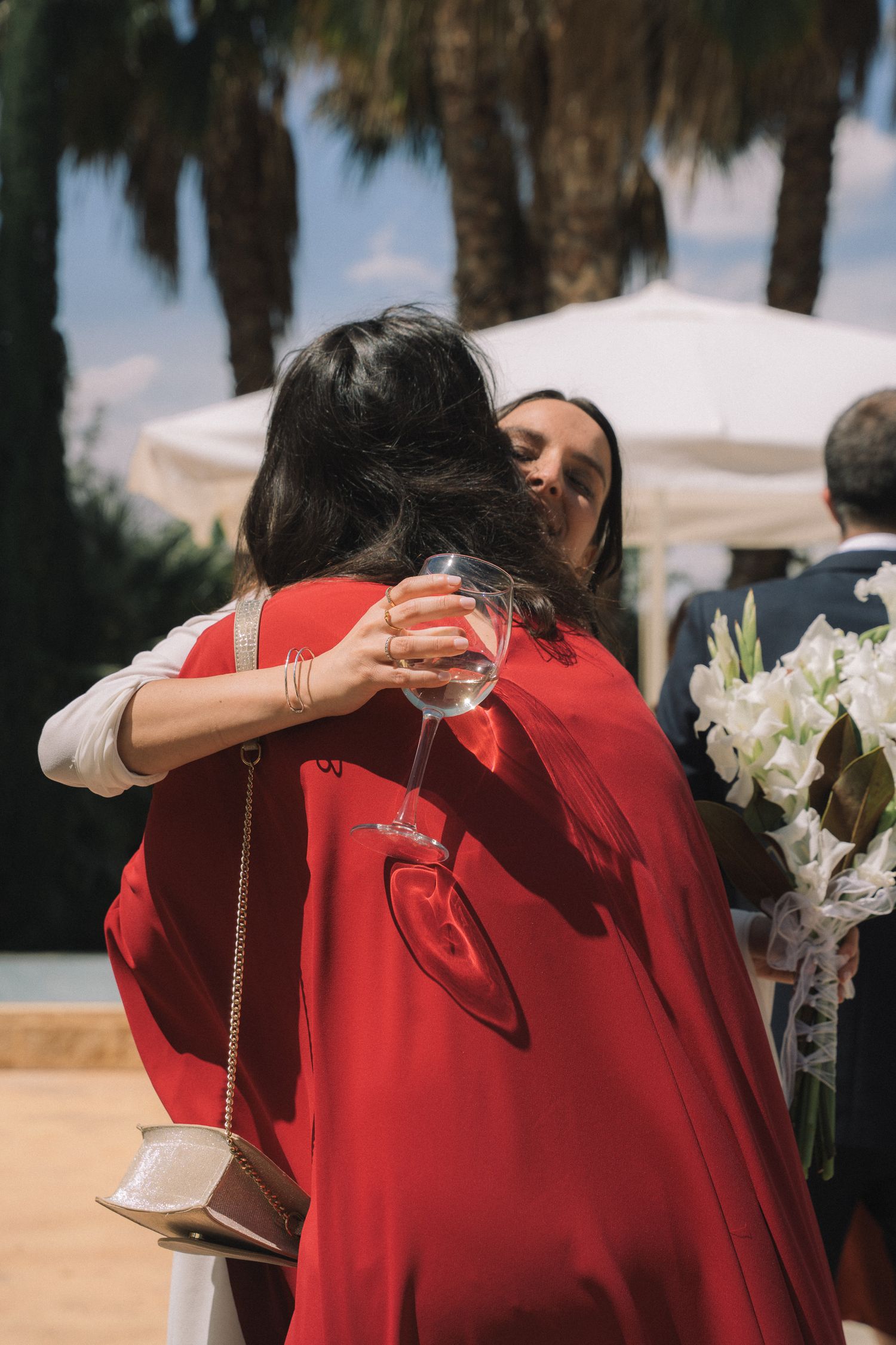 Wedding guests embrace while wearing red dresses as palm trees sway in the background.