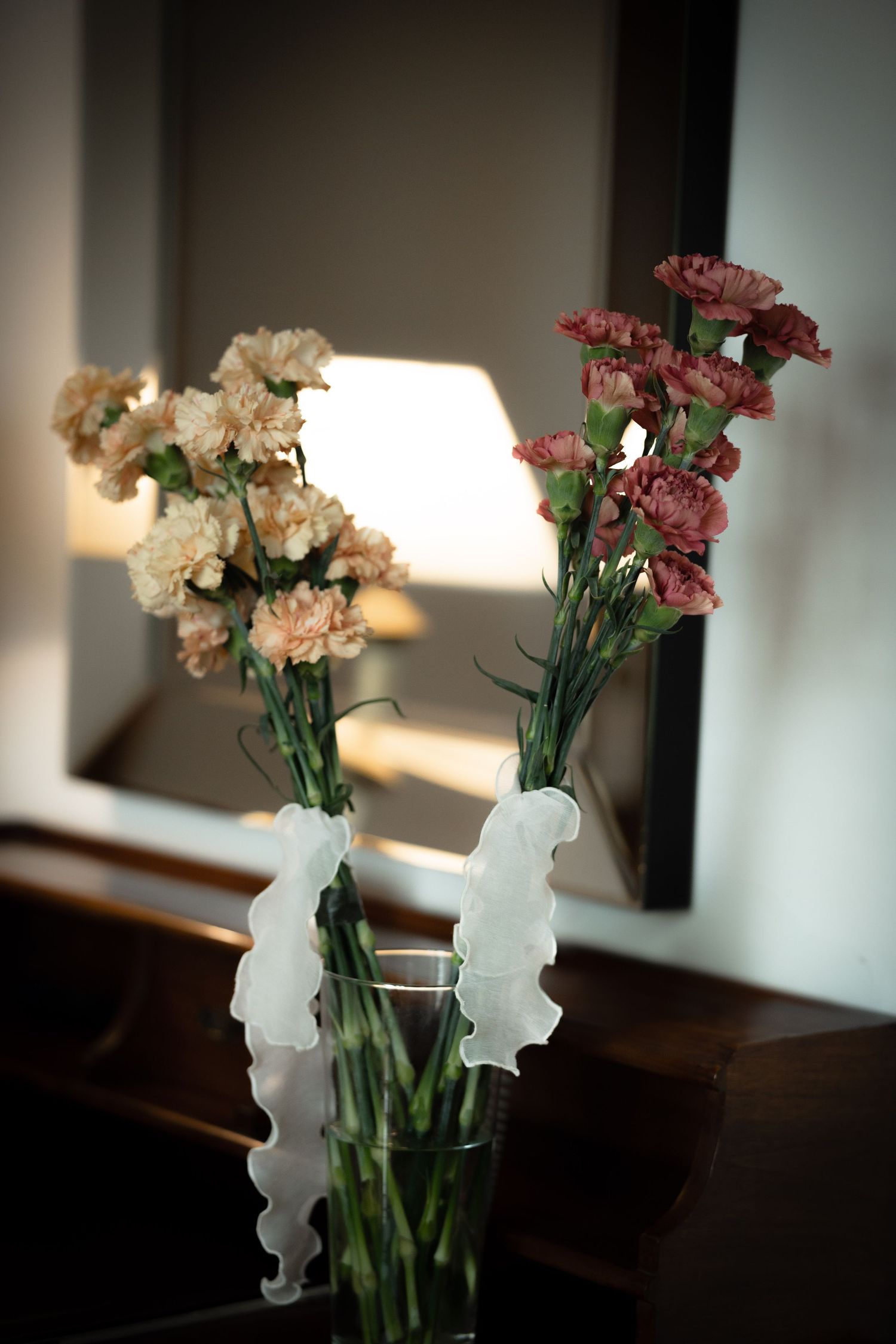 Pink and white carnations in a glass vase with white ribbon decoration.