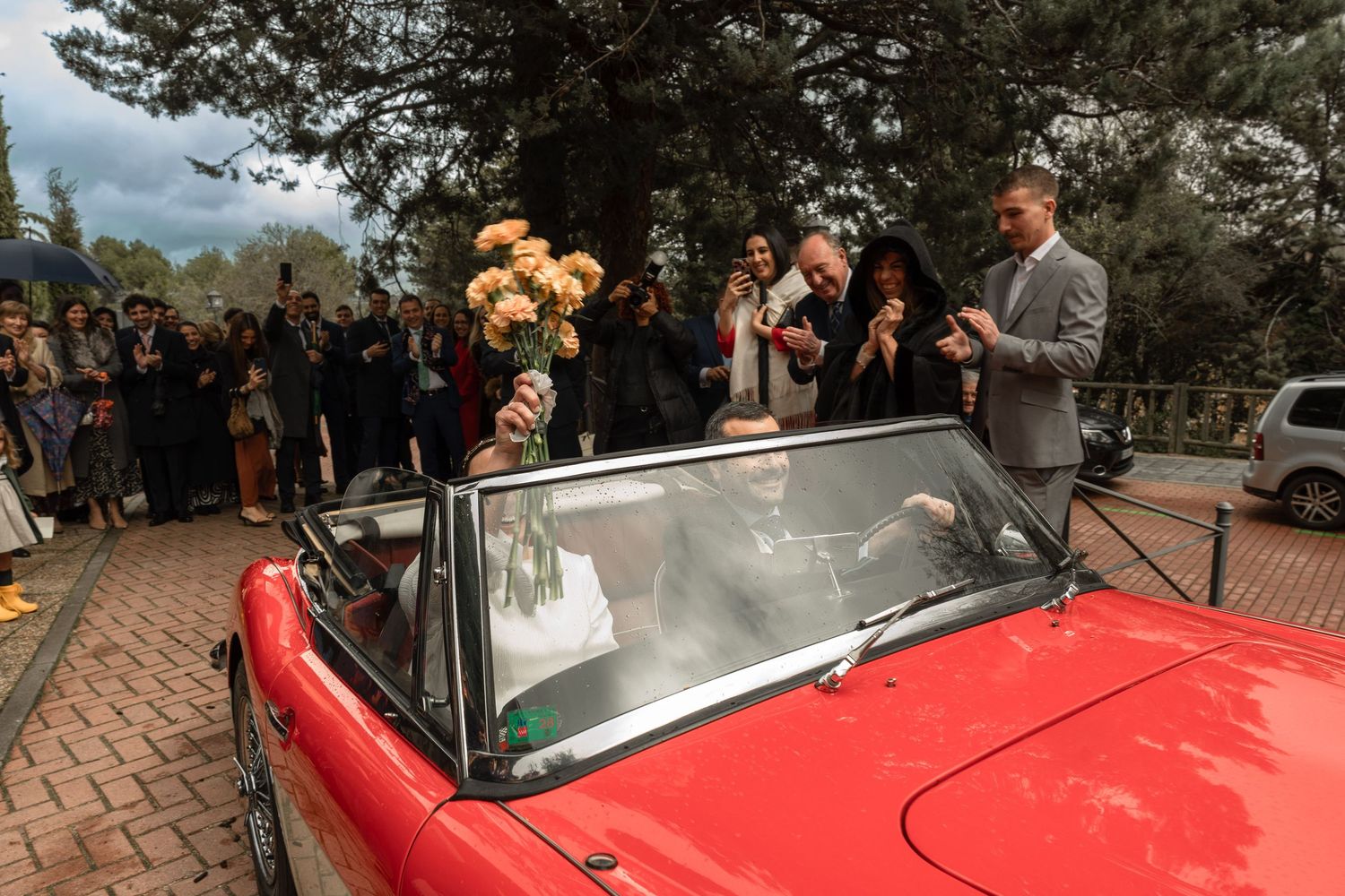 A classic red convertible car with wedding guests gathered around during an outdoor celebration.