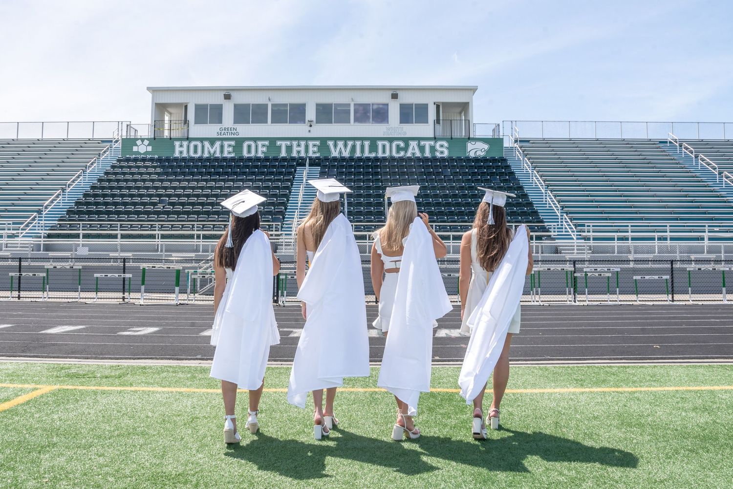 Group of graduates in white gowns pose on a football field with stadium seating in the background.
