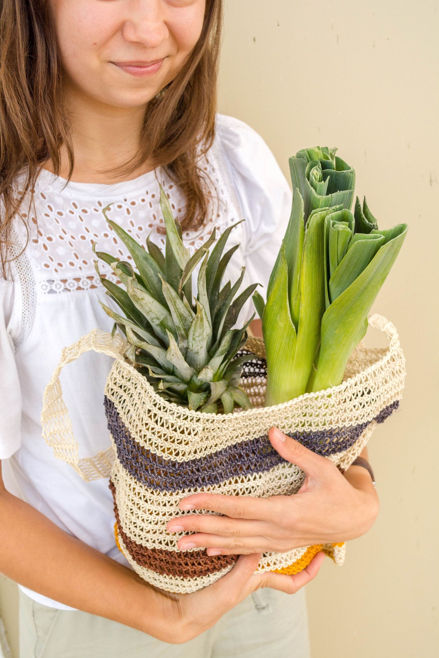A woven basket filled with fresh produce and plants held by someone wearing a white eyelet blouse.