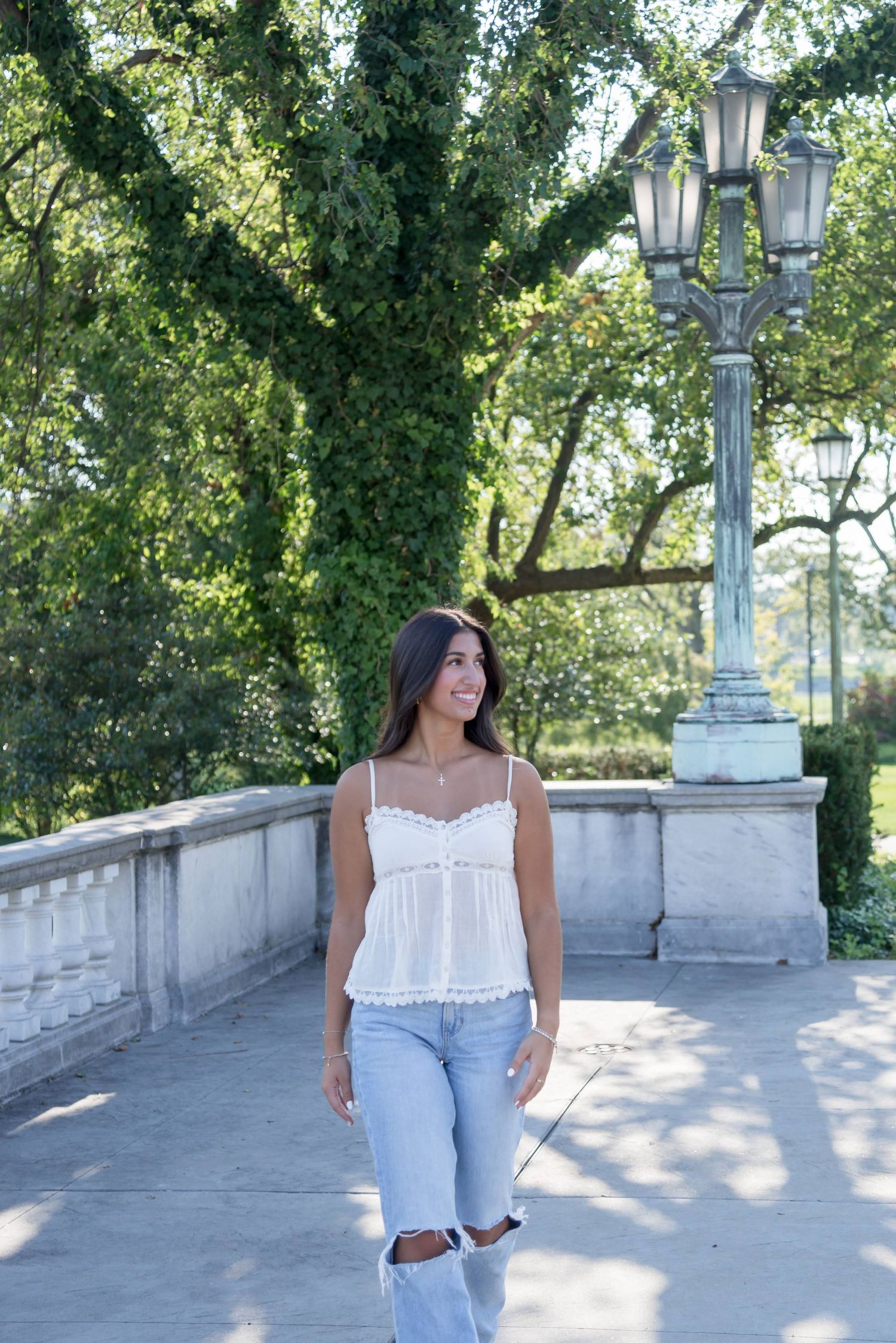 Person in white top and jeans poses along a stone balcony with ornate lampposts and lush greenery.