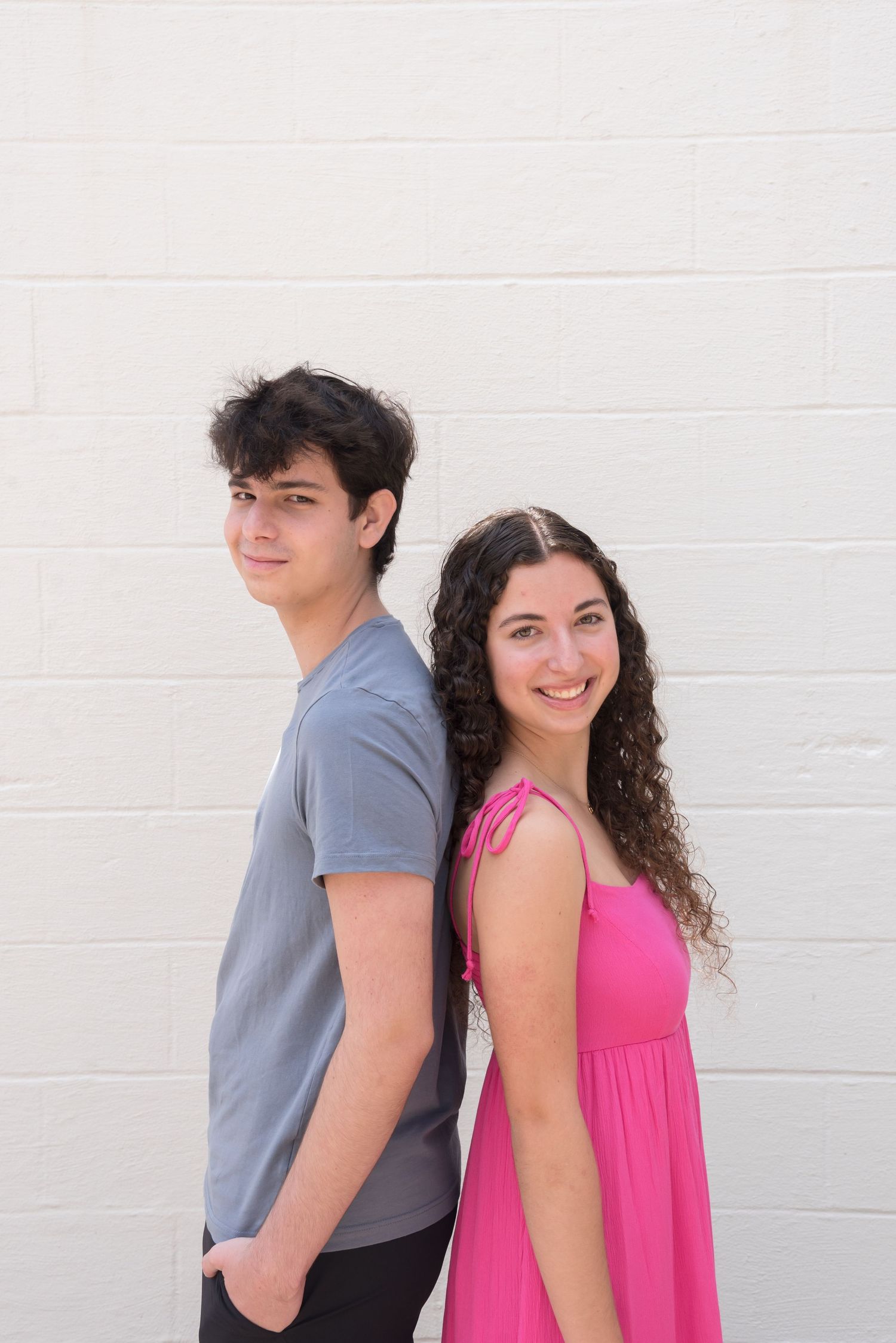 A couple poses against a white wall, with one wearing a pink top and the other in gray.