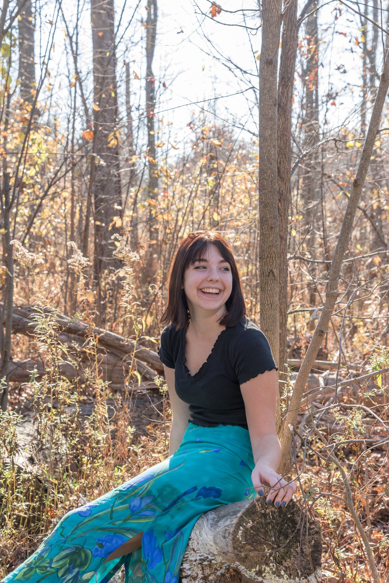 Person in teal skirt and black top poses playfully in autumn woods setting.