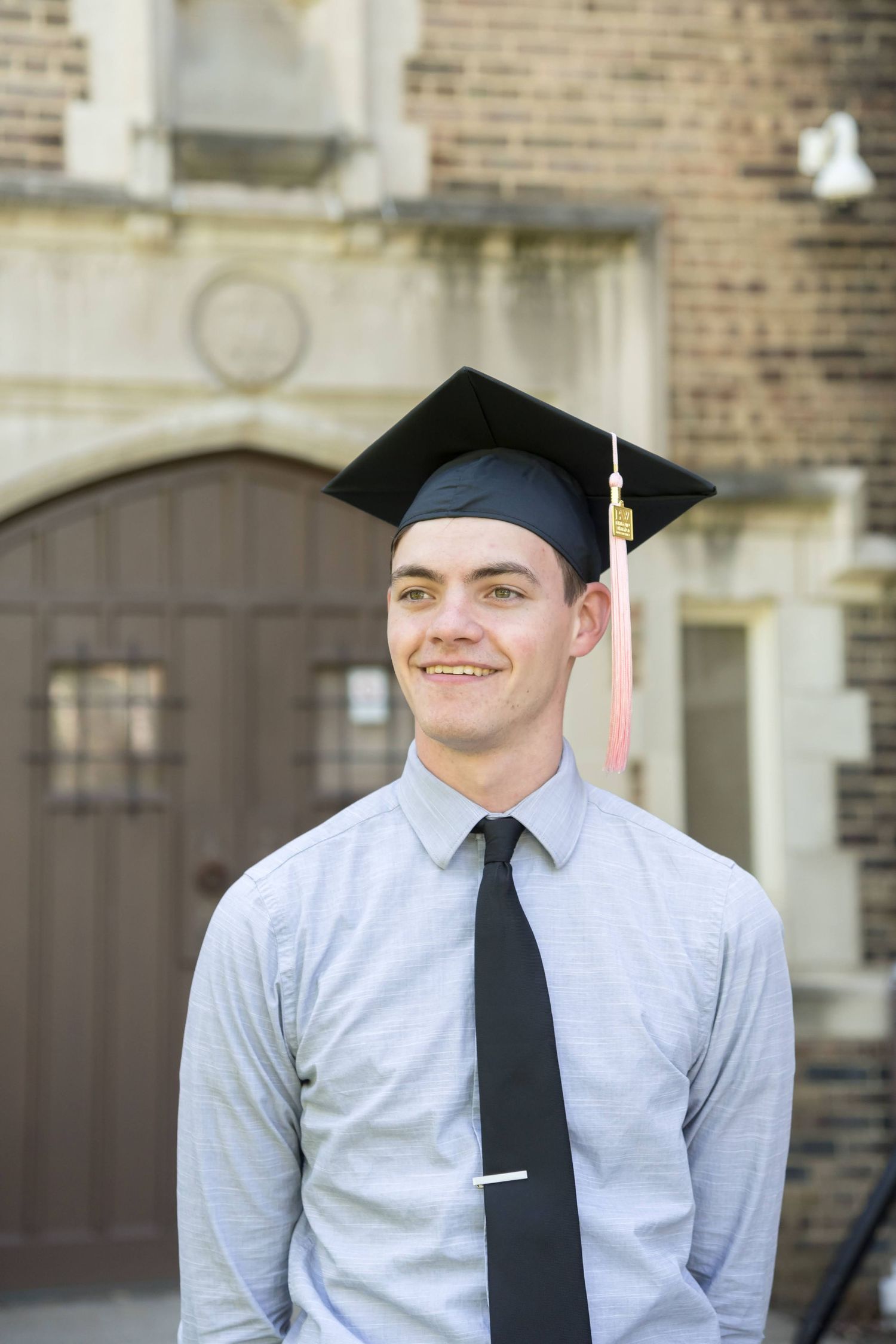 Graduate wearing cap and gown poses in front of brick building.