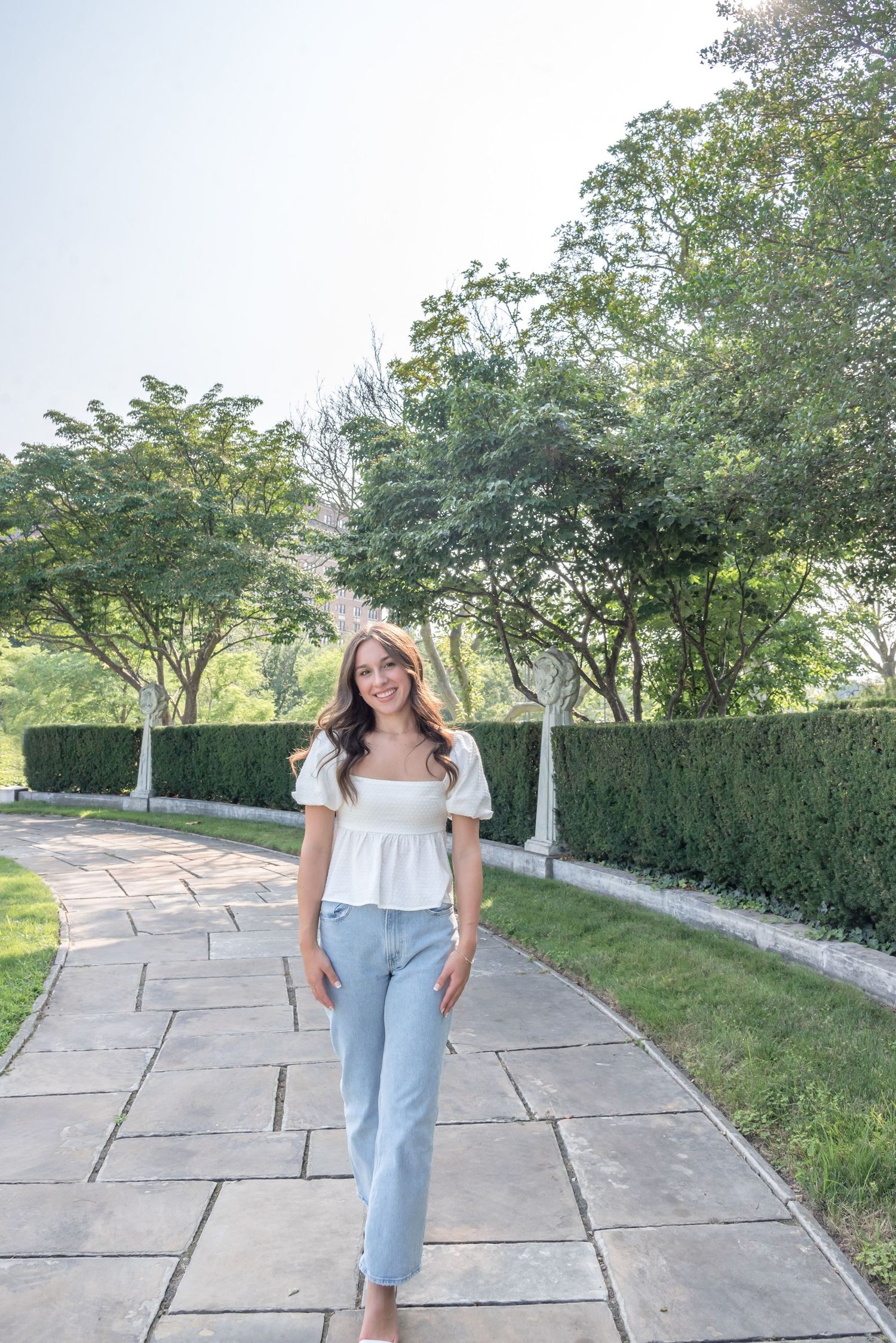 Person in white top and jeans walks along stone path bordered by manicured hedges.