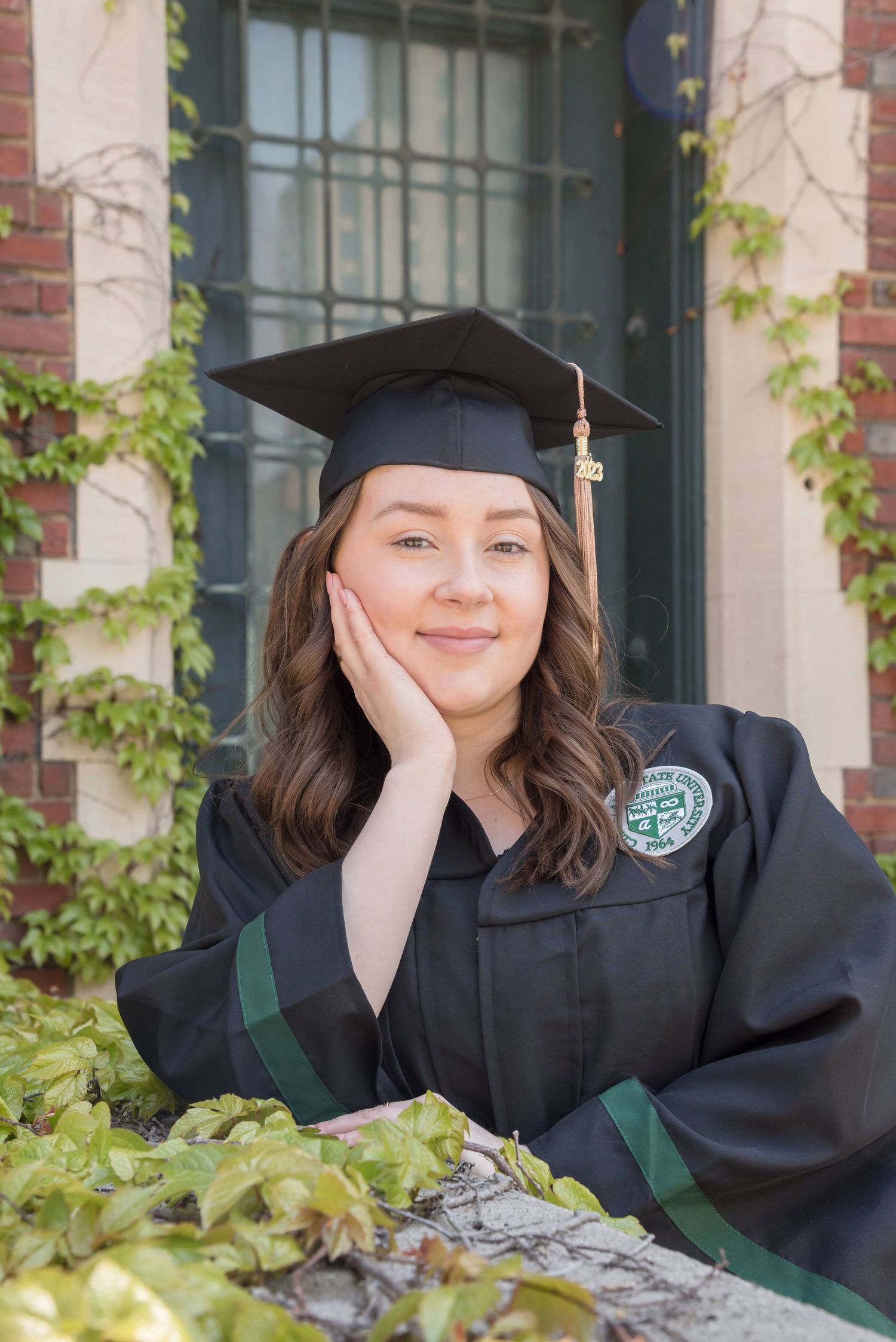 Graduate in cap and gown posing for photos against ivy-covered brick wall.