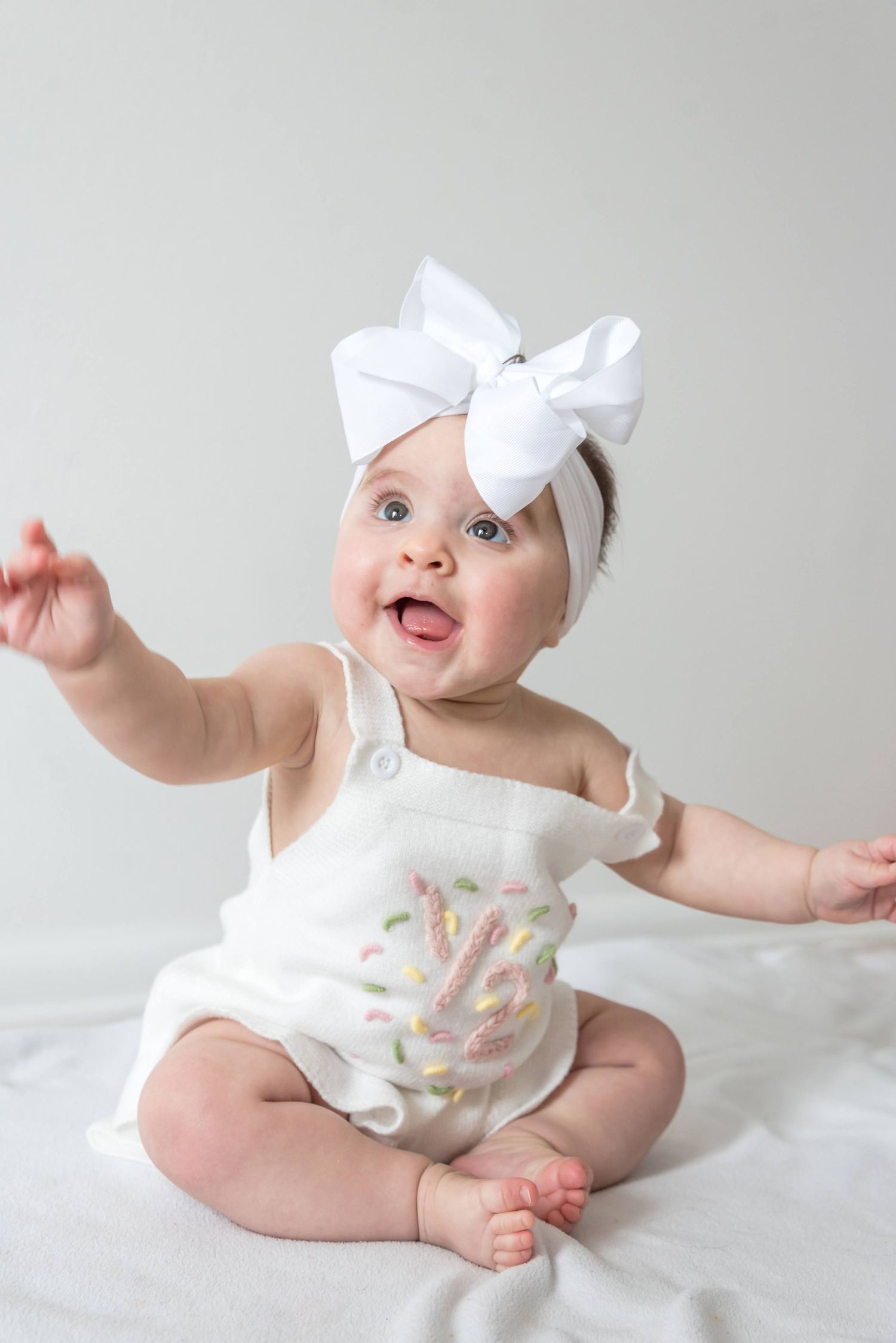 Baby in white outfit with bow headband smiling and reaching out.