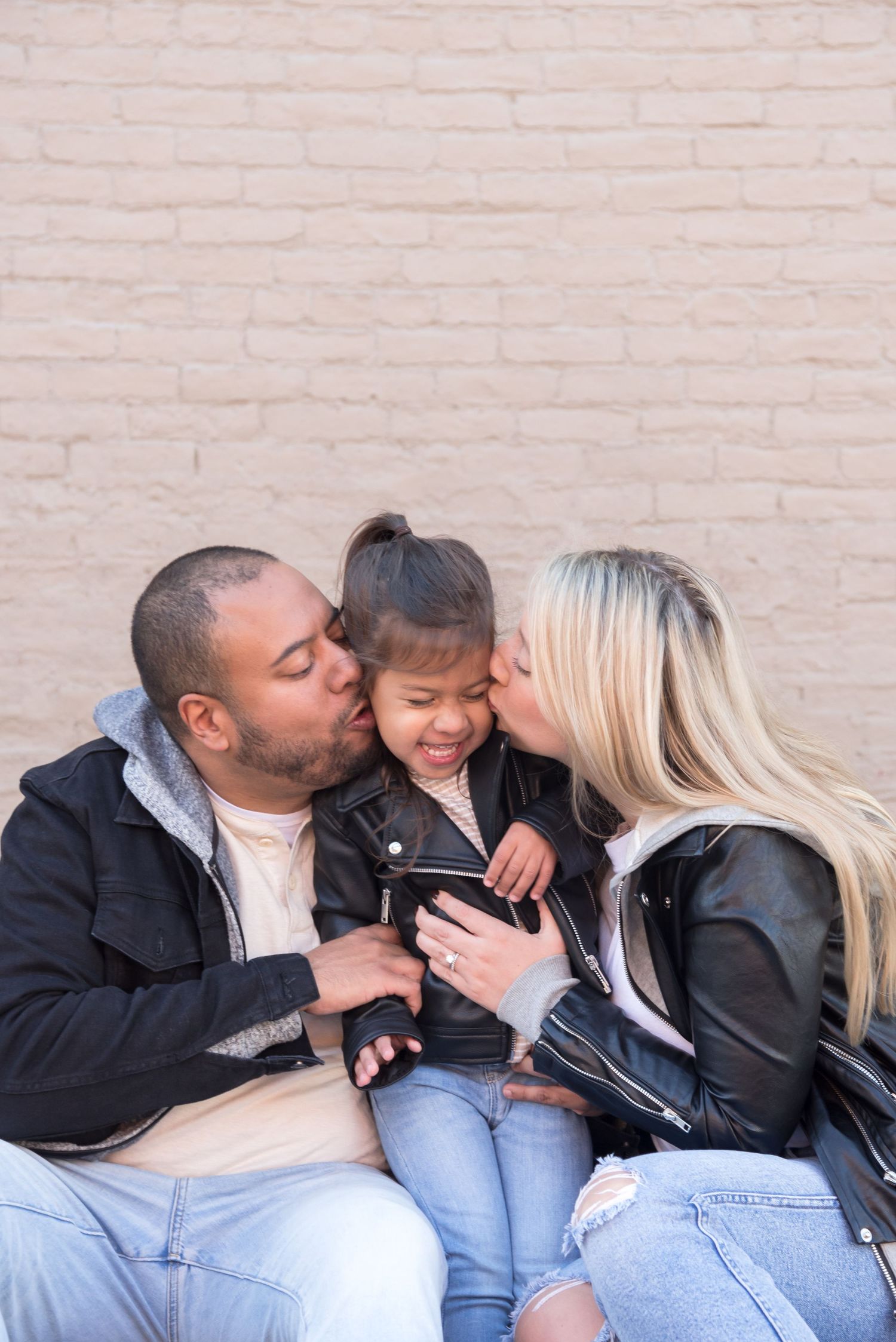 Family sharing affectionate moment together against light colored brick wall.