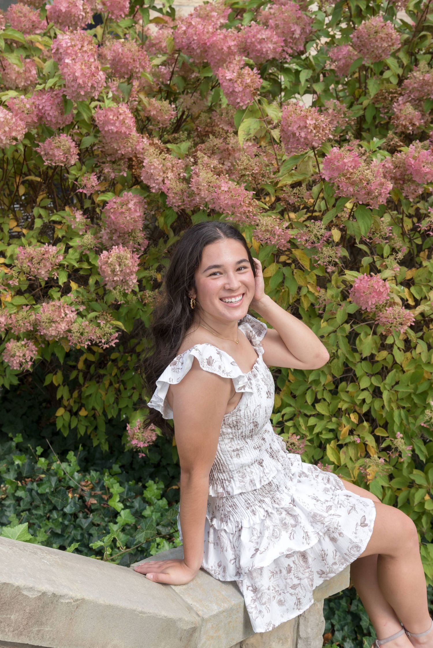 Person in white floral dress poses by blooming pink flowers on stone ledge.