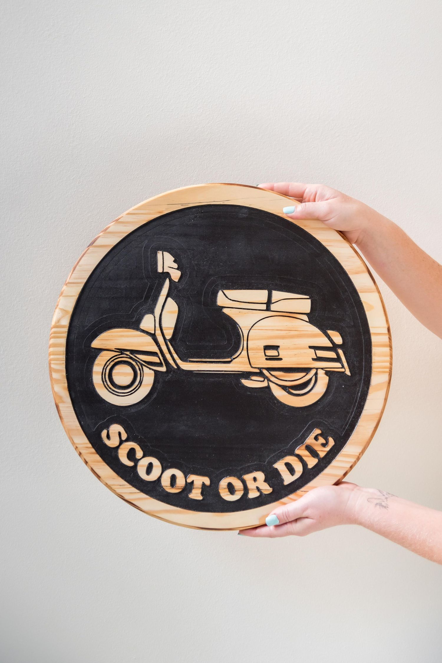 Wooden sign with vintage scooter design and text 'Scoot or Die' being held against white background.