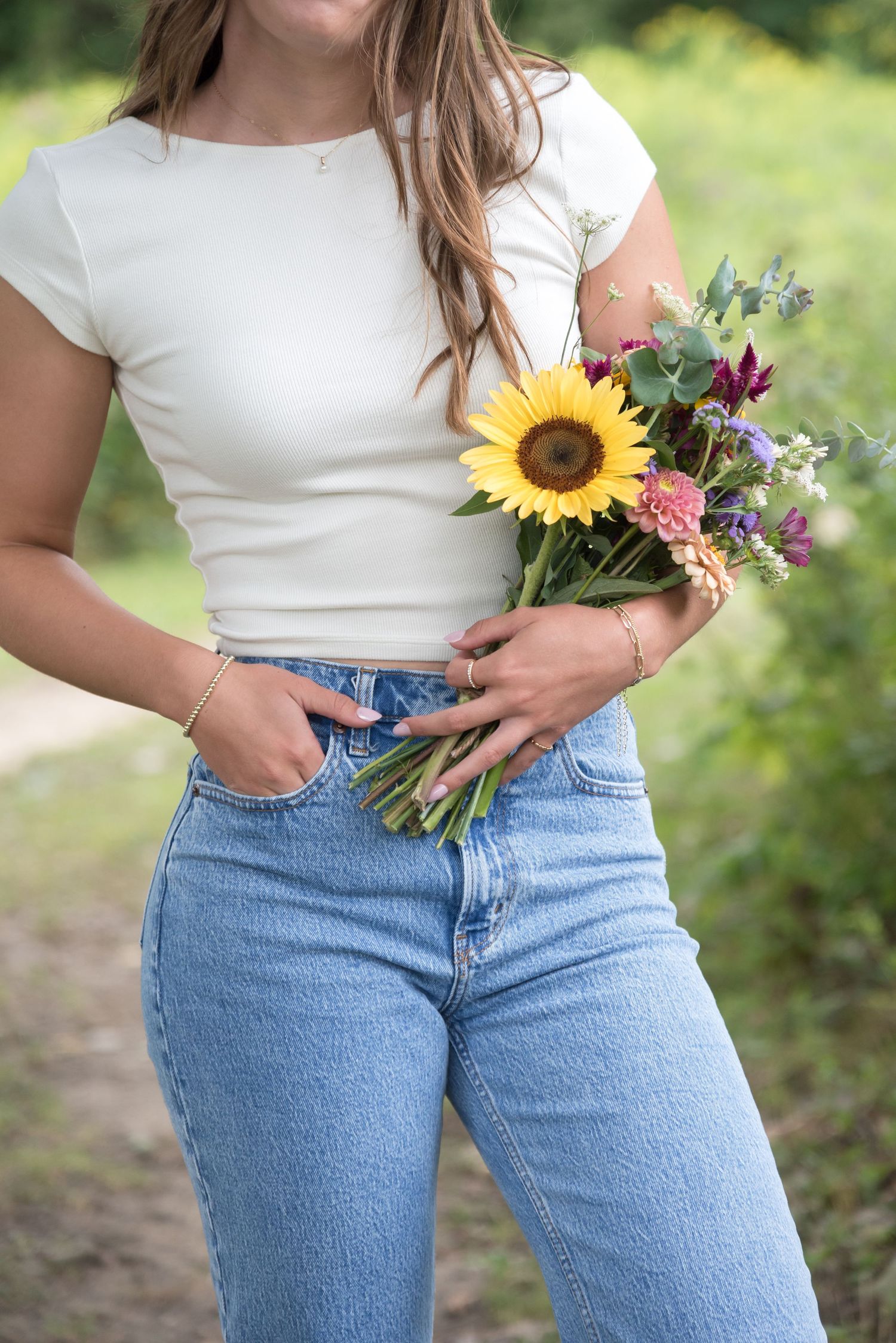 Woman in white top and jeans holding sunflowers and wildflowers against a green background.