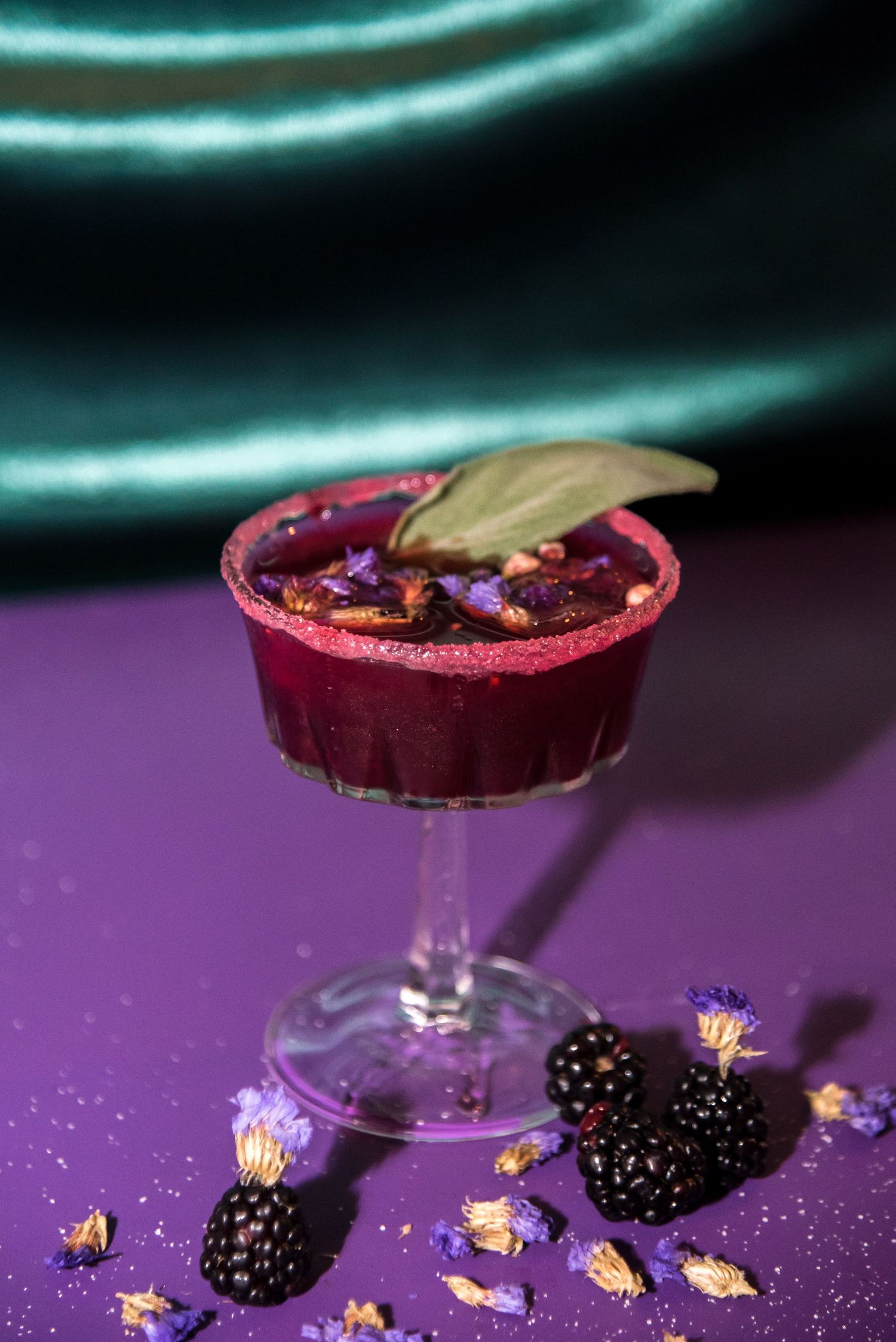 Elegant cocktail glass with blackberries and edible flowers on a purple background.