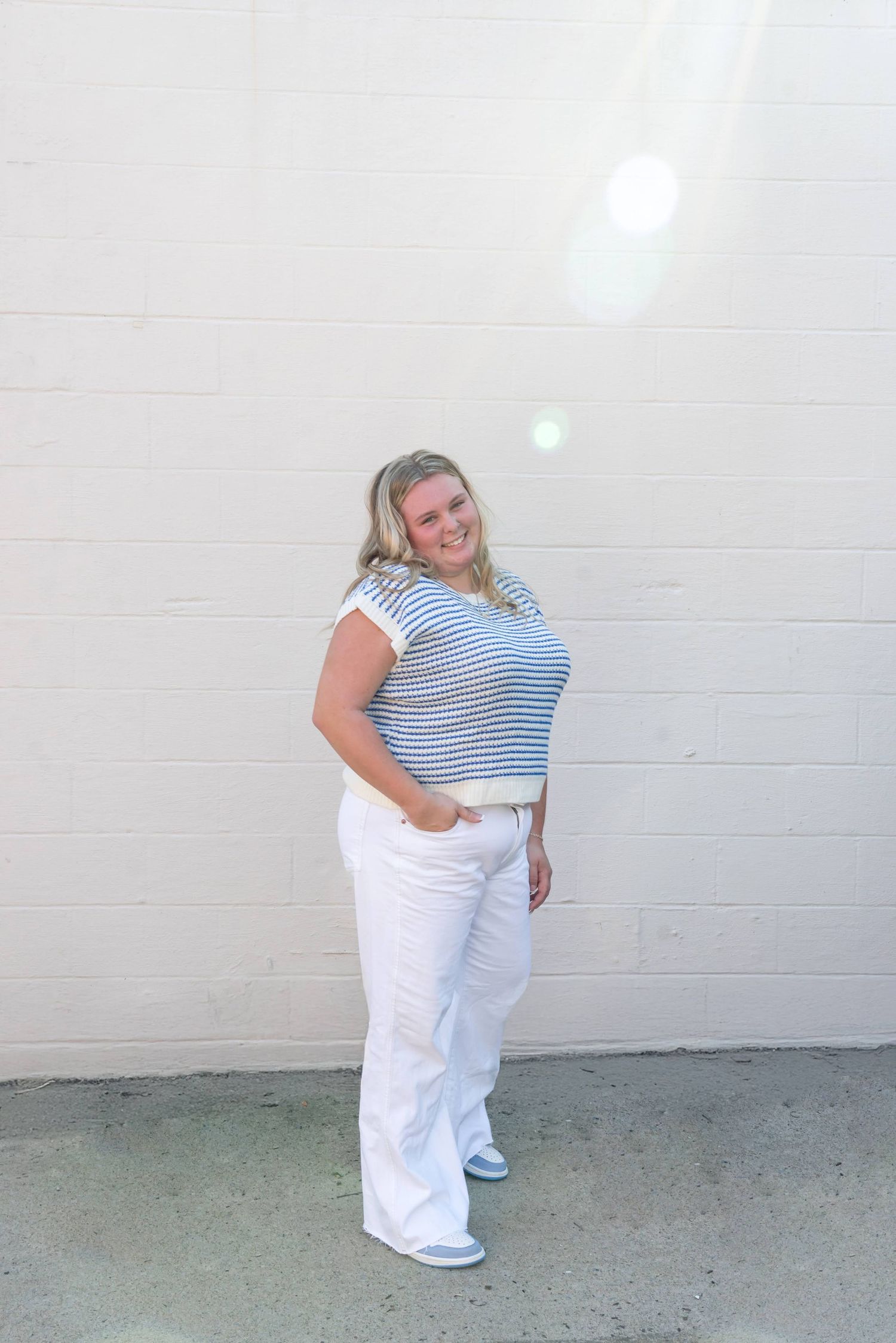 Person in striped top and white pants poses casually against white wall.
