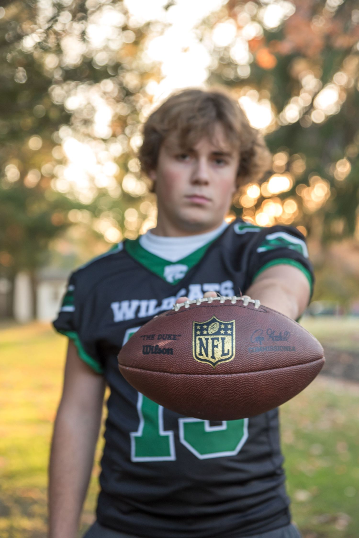 A football player in a green jersey holds an NFL football in golden sunset light.
