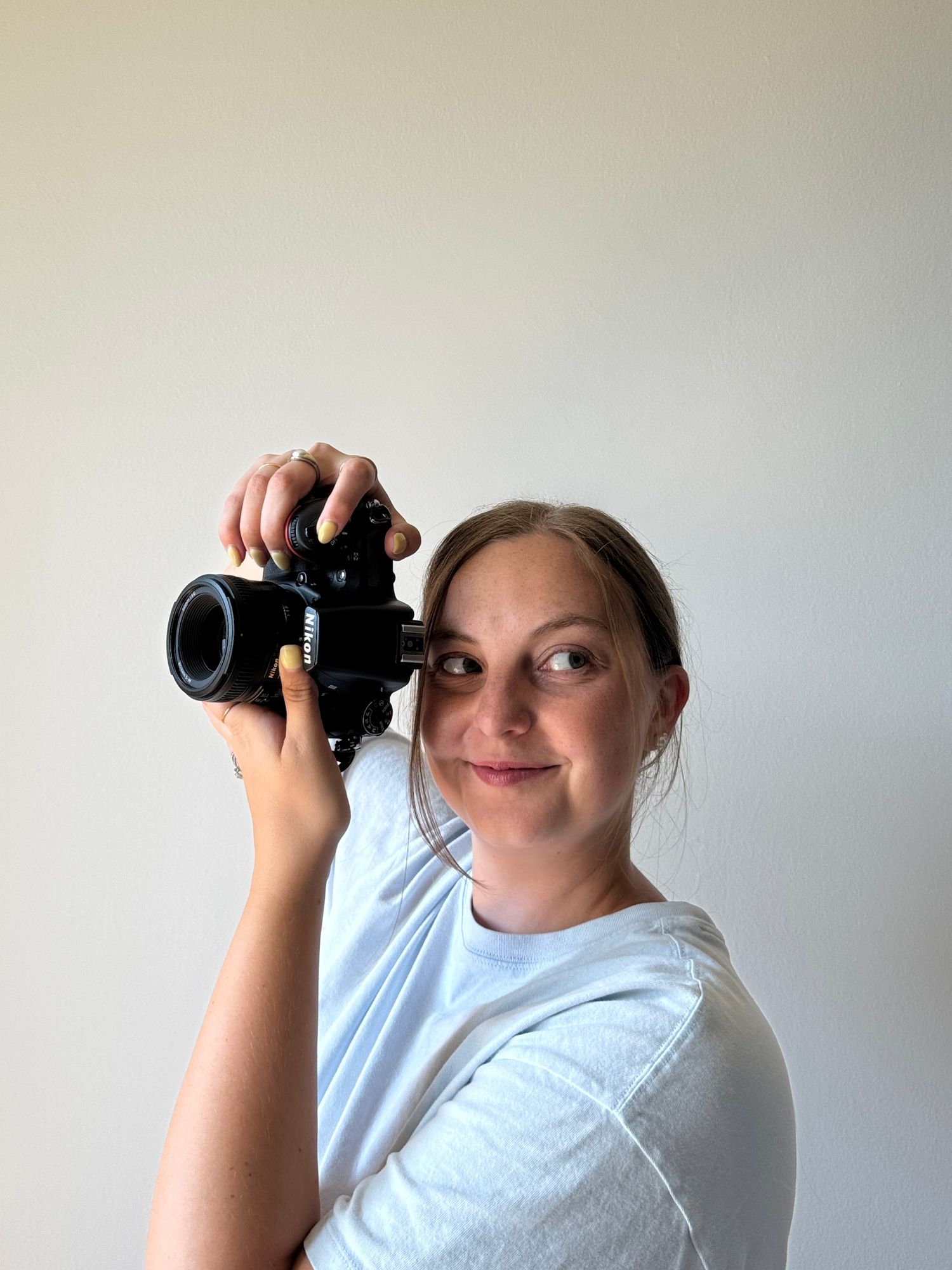 Young woman in white t-shirt holding a black DSLR camera up to her face, smiling against a white background.