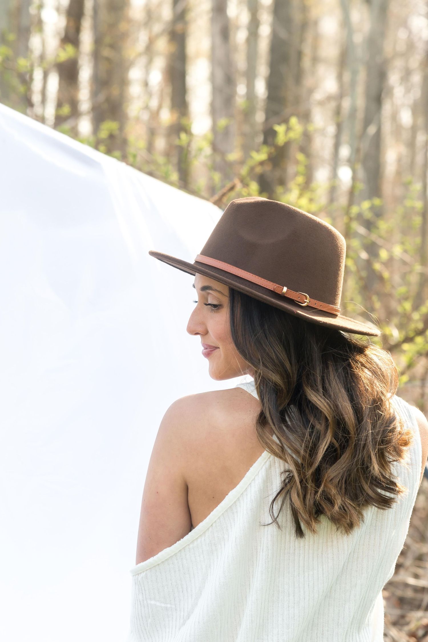 Woman in white sweater and brown hat poses against natural forest backdrop.