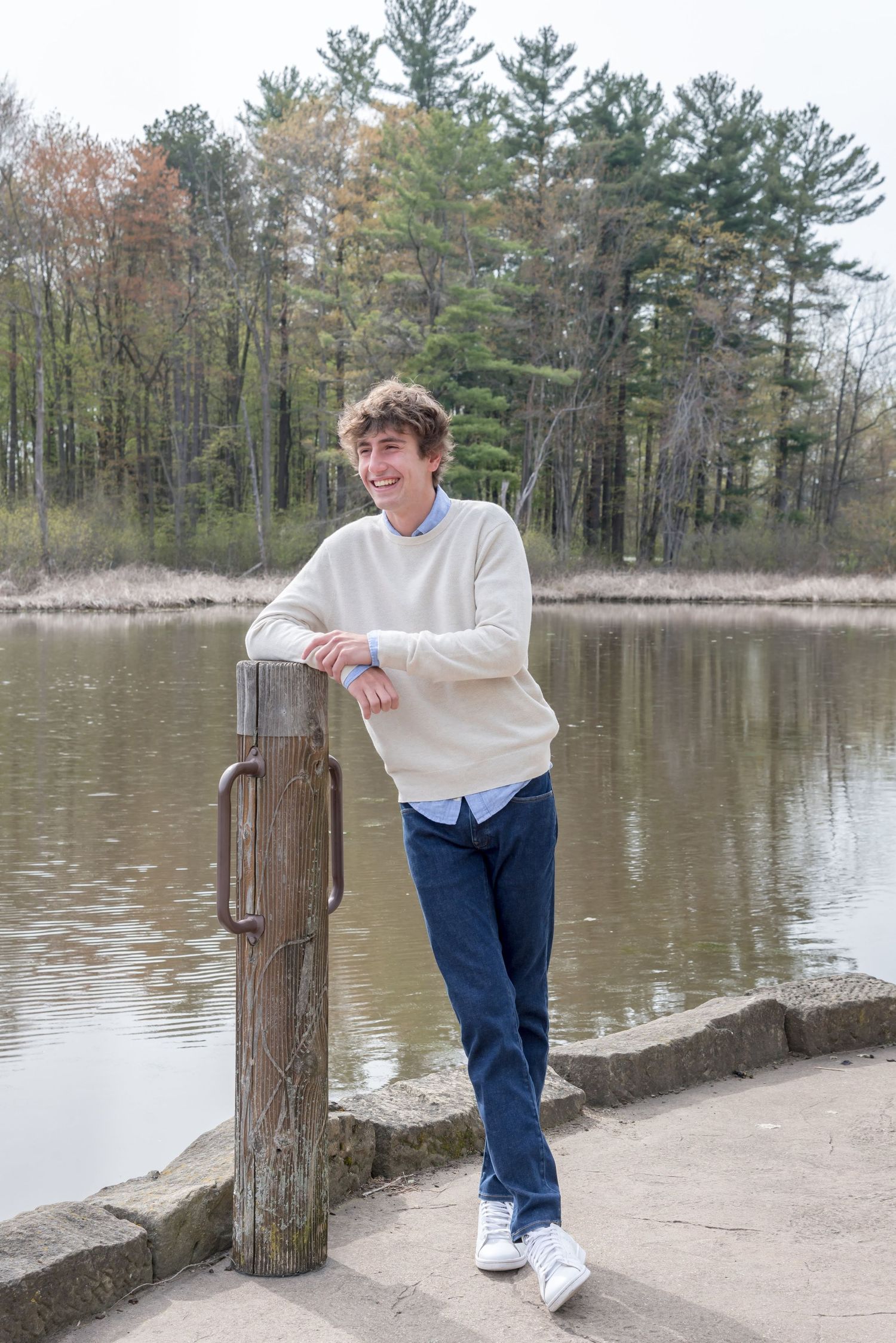 Person in cream sweater leans on wooden post by serene lake.