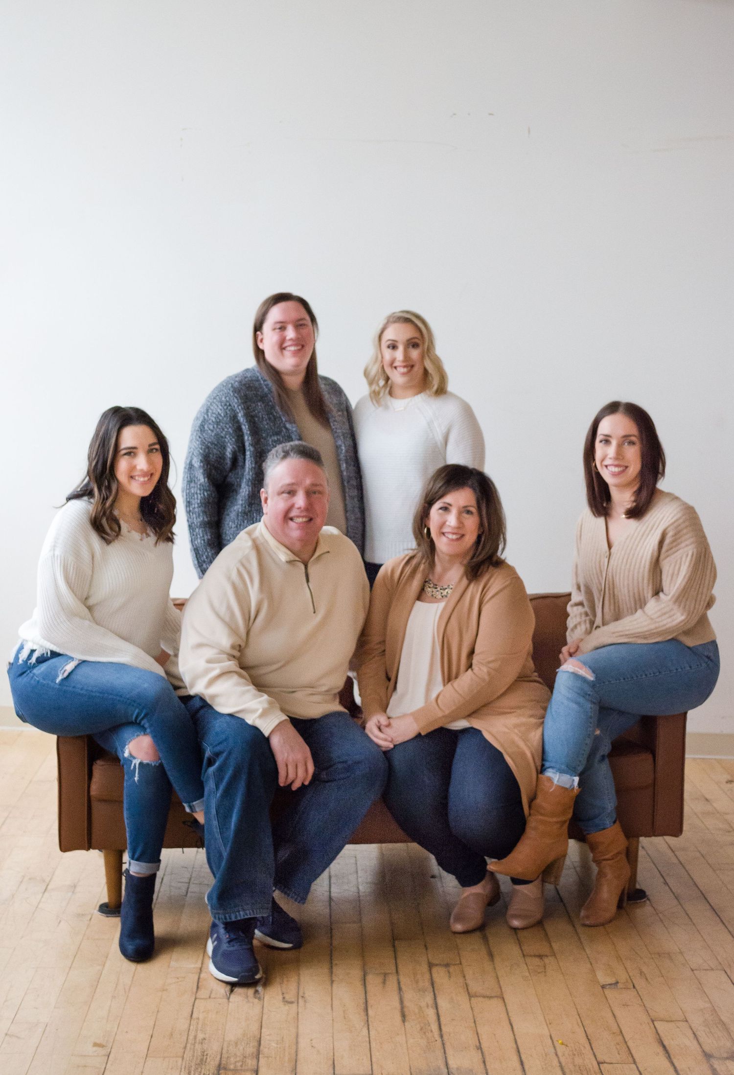 A group portrait of six people arranged casually on a brown couch against a white wall.