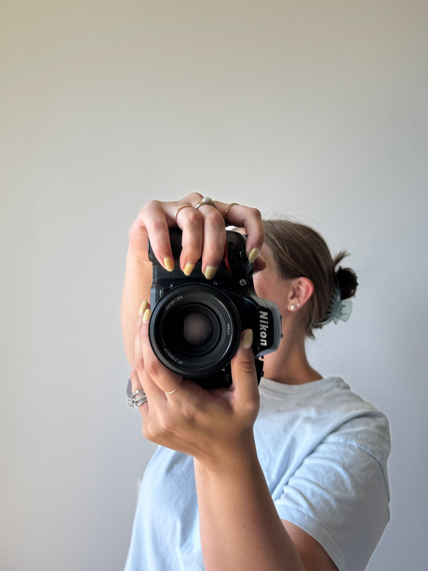 Woman in white t-shirt holding a Nikon DSLR camera up to her face, wearing rings and green nail polish.