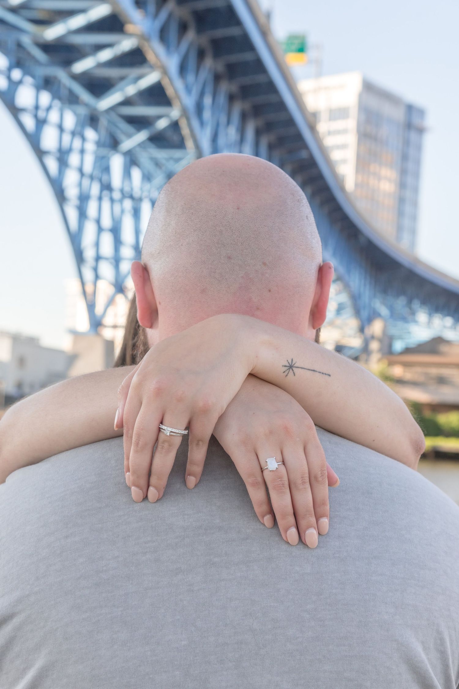 Close-up of hands with wedding rings embracing against bridge backdrop.