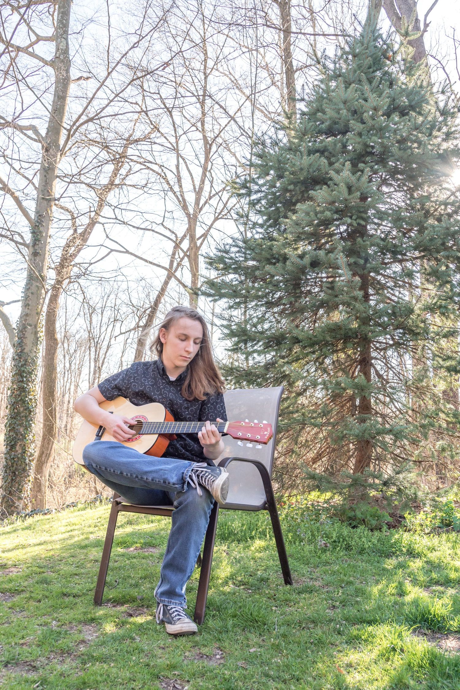 Person playing acoustic guitar while sitting on grassy hill surrounded by trees.
