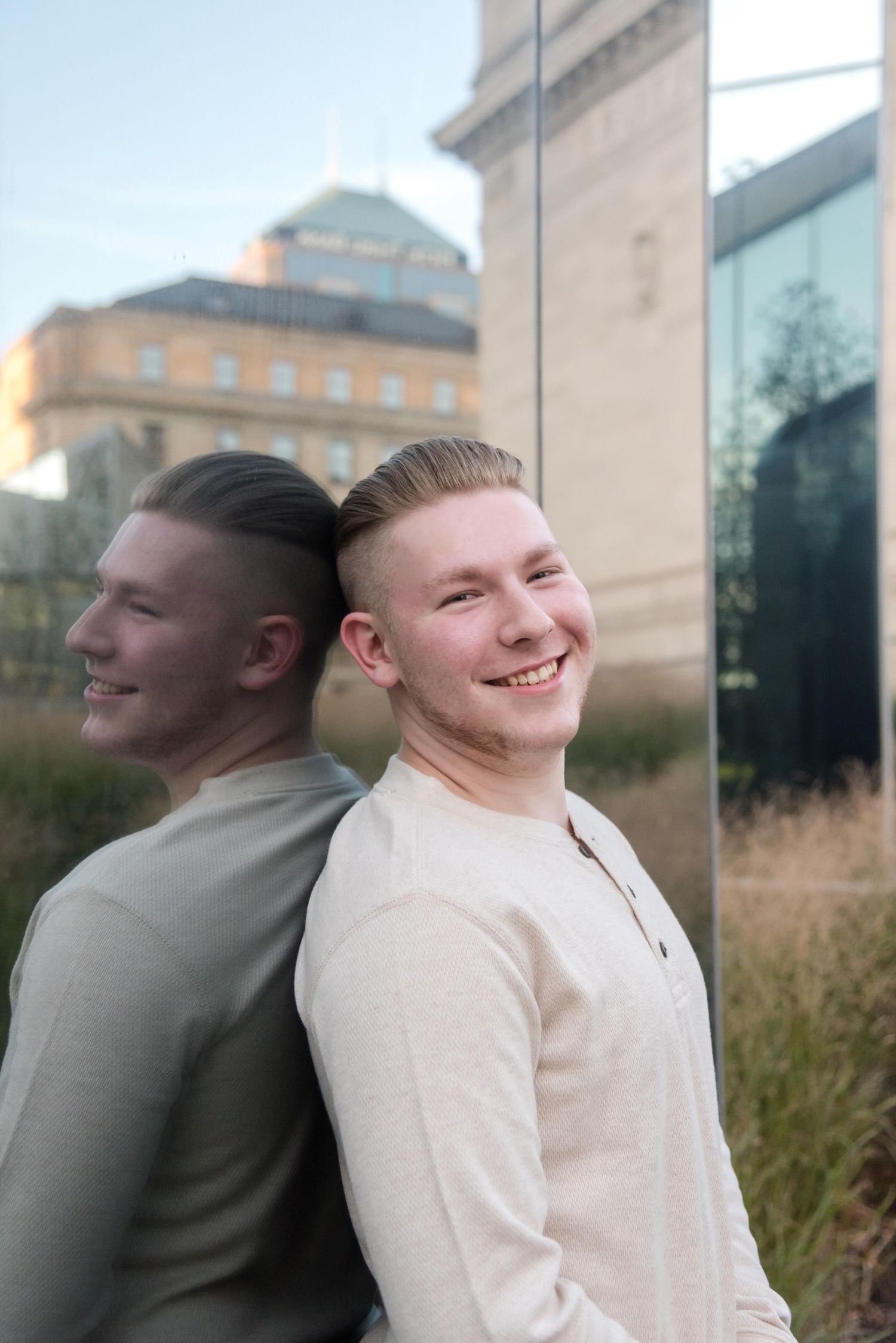 Person in white sweater leans against reflective glass building wall at sunset.