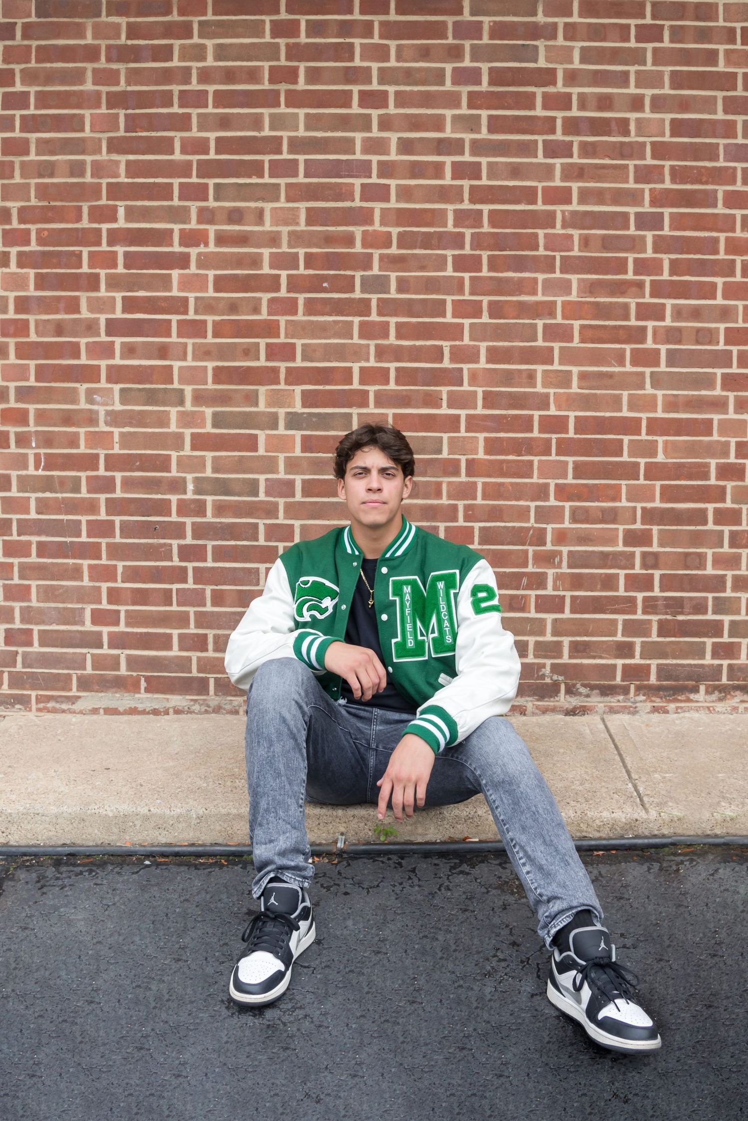 Student in varsity jacket sitting against brick wall wearing sneakers.