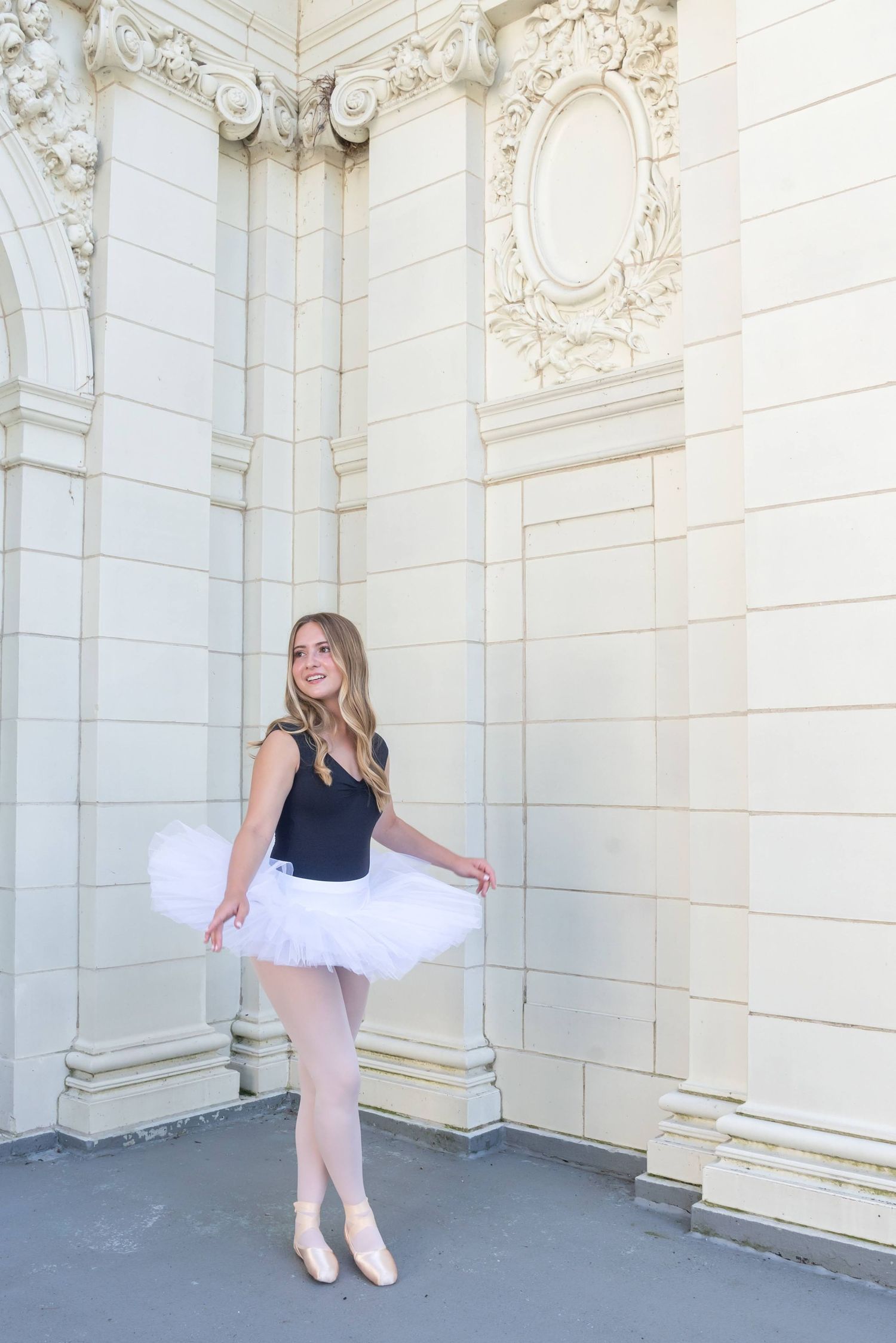 Dancer in white tutu and black leotard poses against ornate white stone wall.