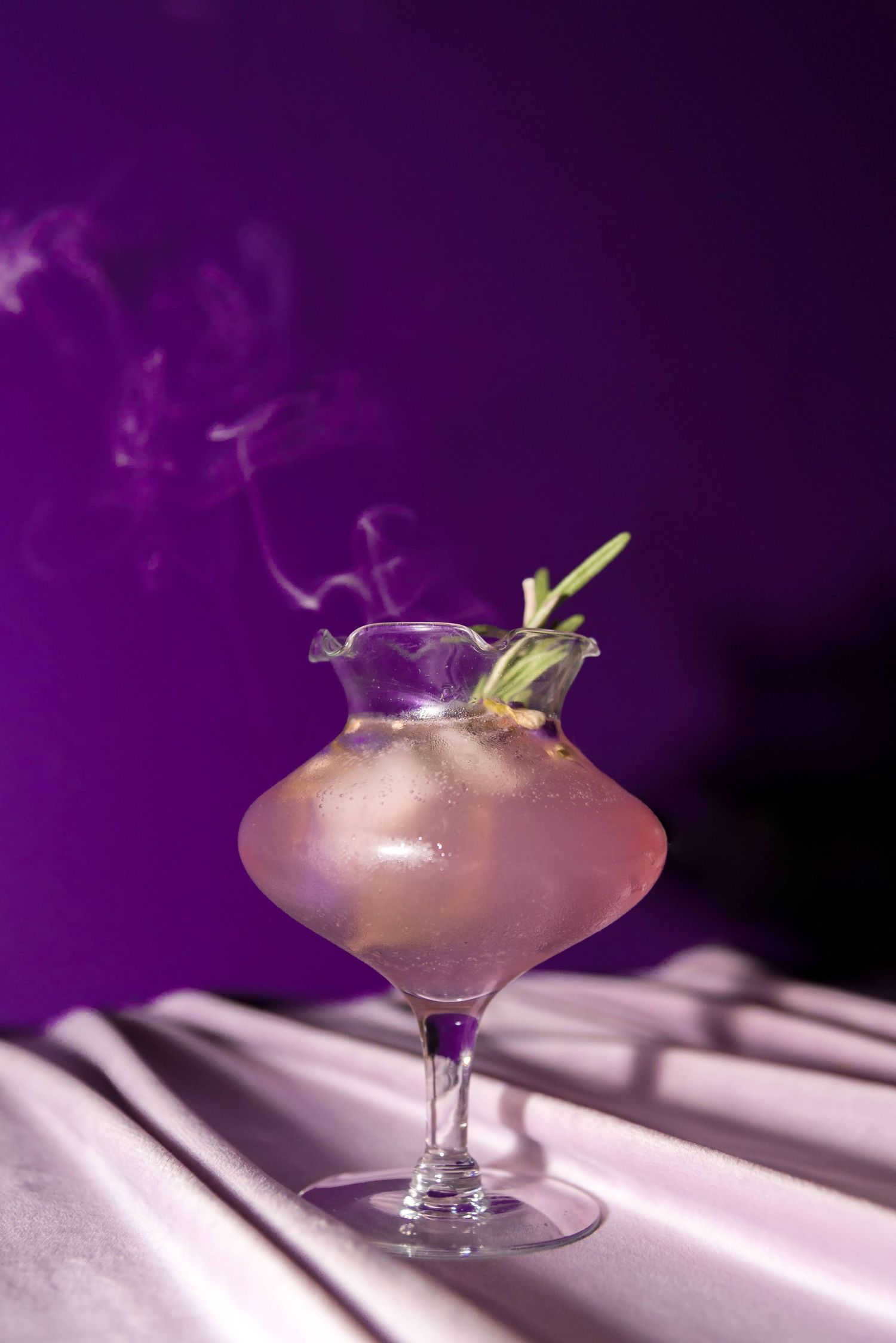 Pink cocktail with garnish in coupe glass against dramatic purple lighting.