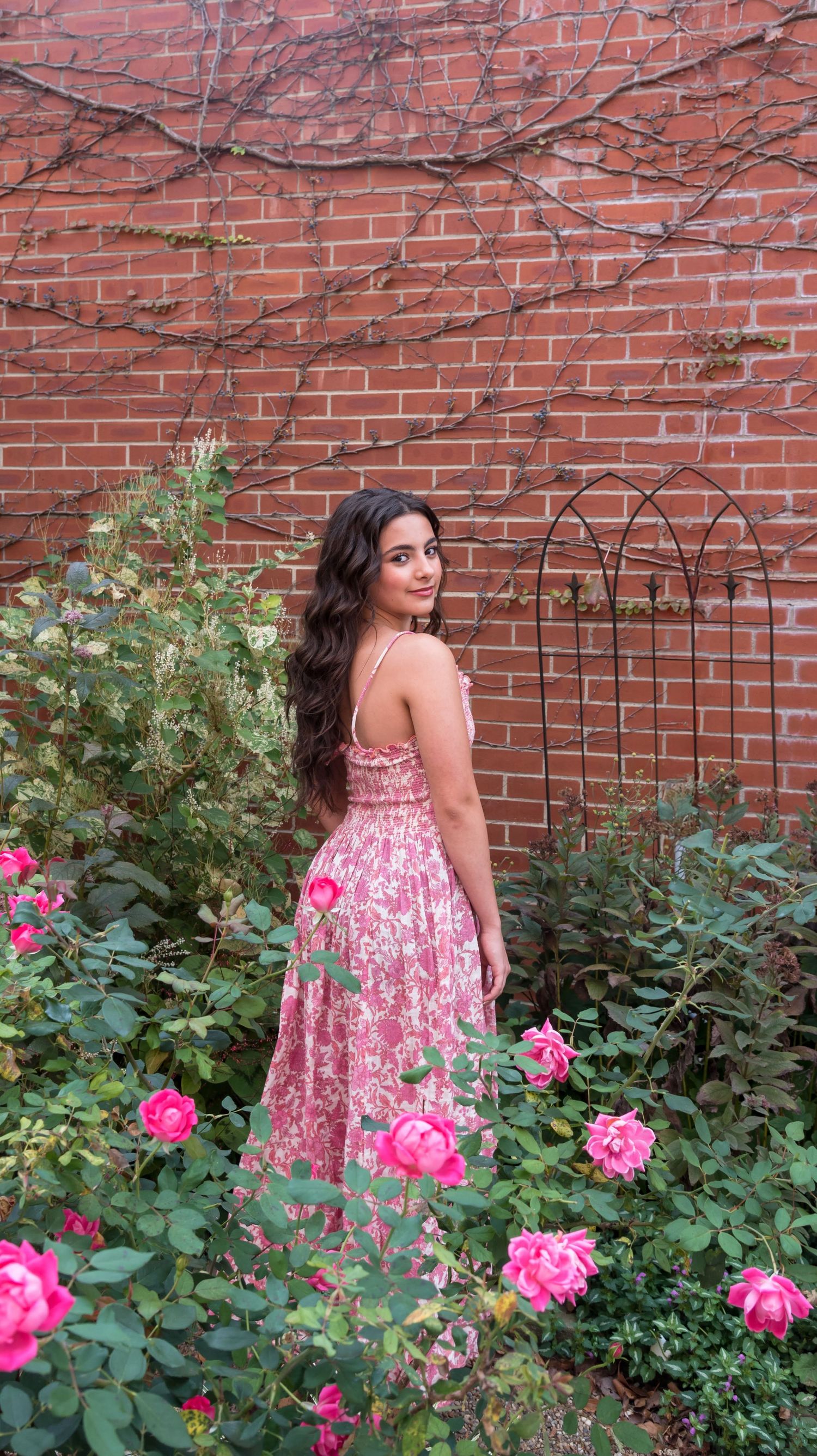 Person in floral pink dress poses by brick wall with blooming rose garden.