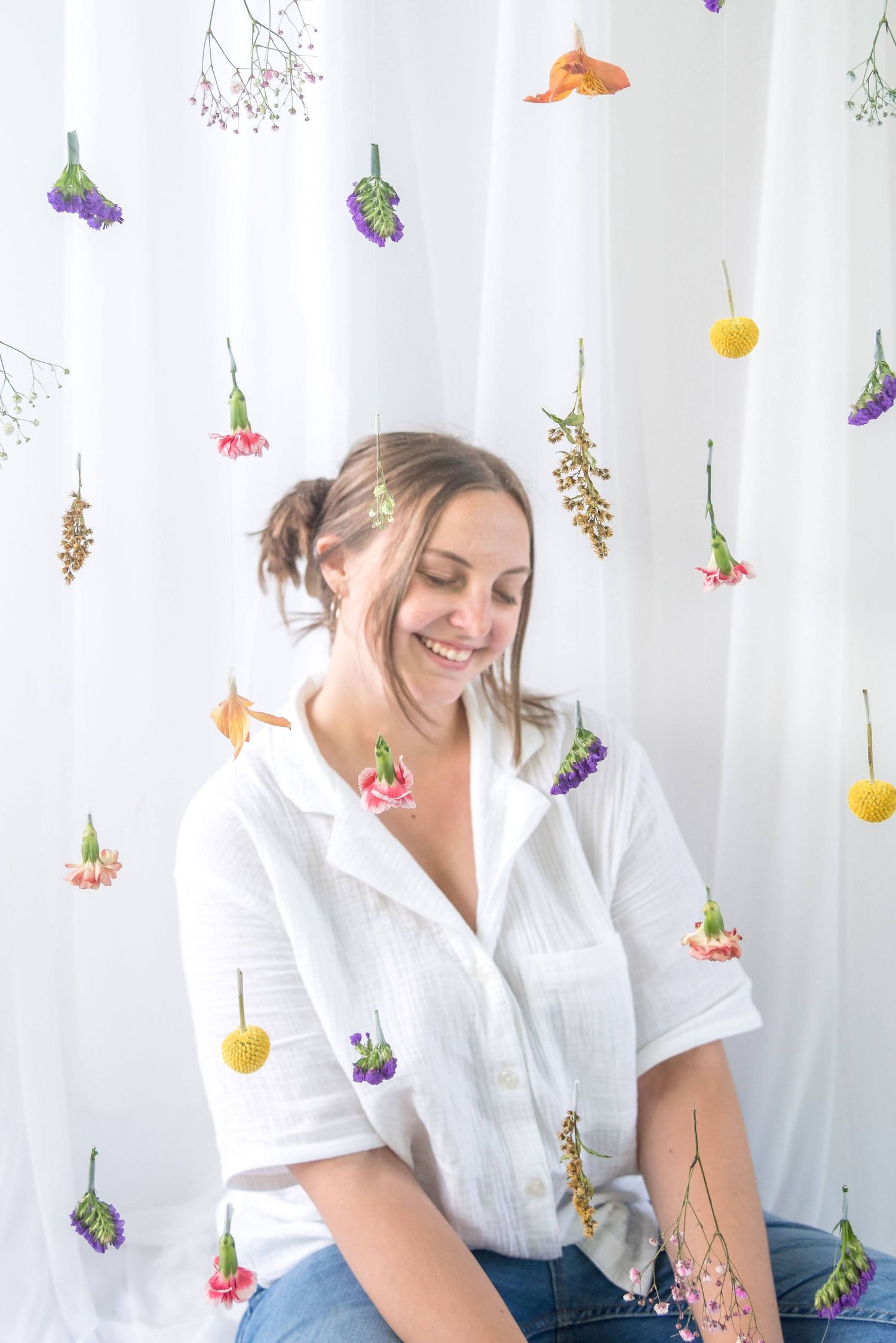 Woman in white shirt poses playfully against white curtain with hanging dried flowers.