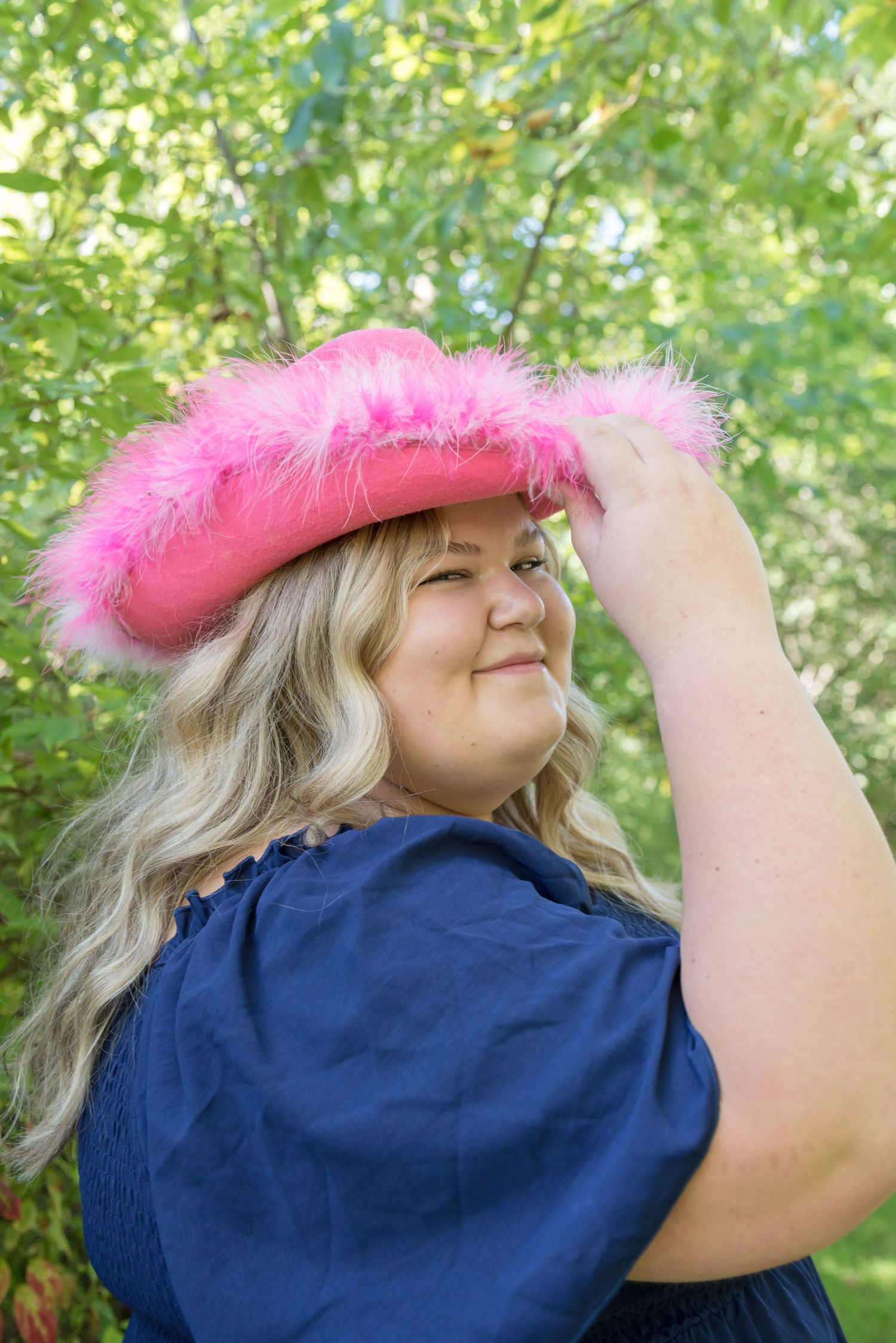 Person wearing a fluffy pink hat poses playfully against green foliage.