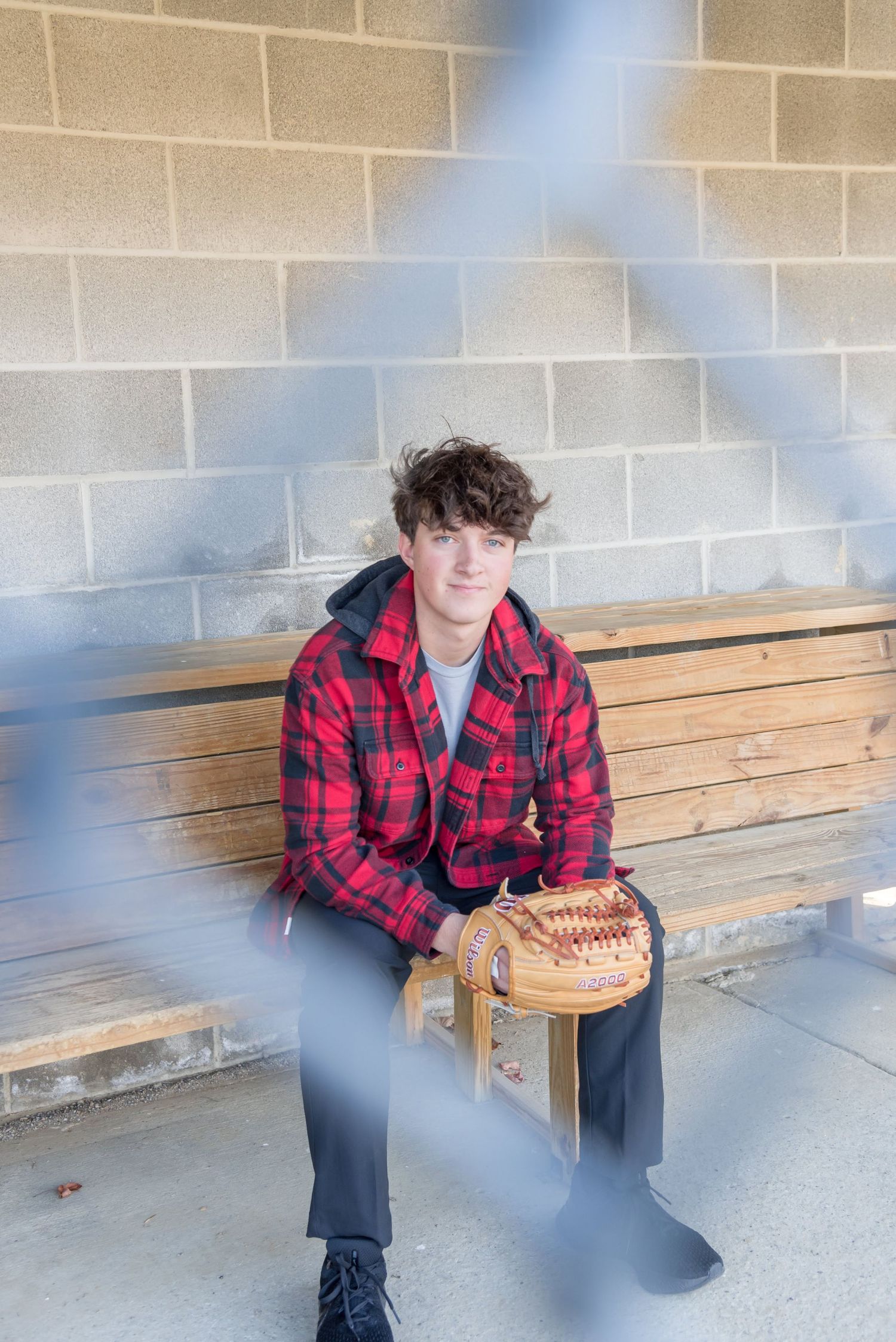 Baseball player in red plaid flannel sits on wooden bench with baseball glove.