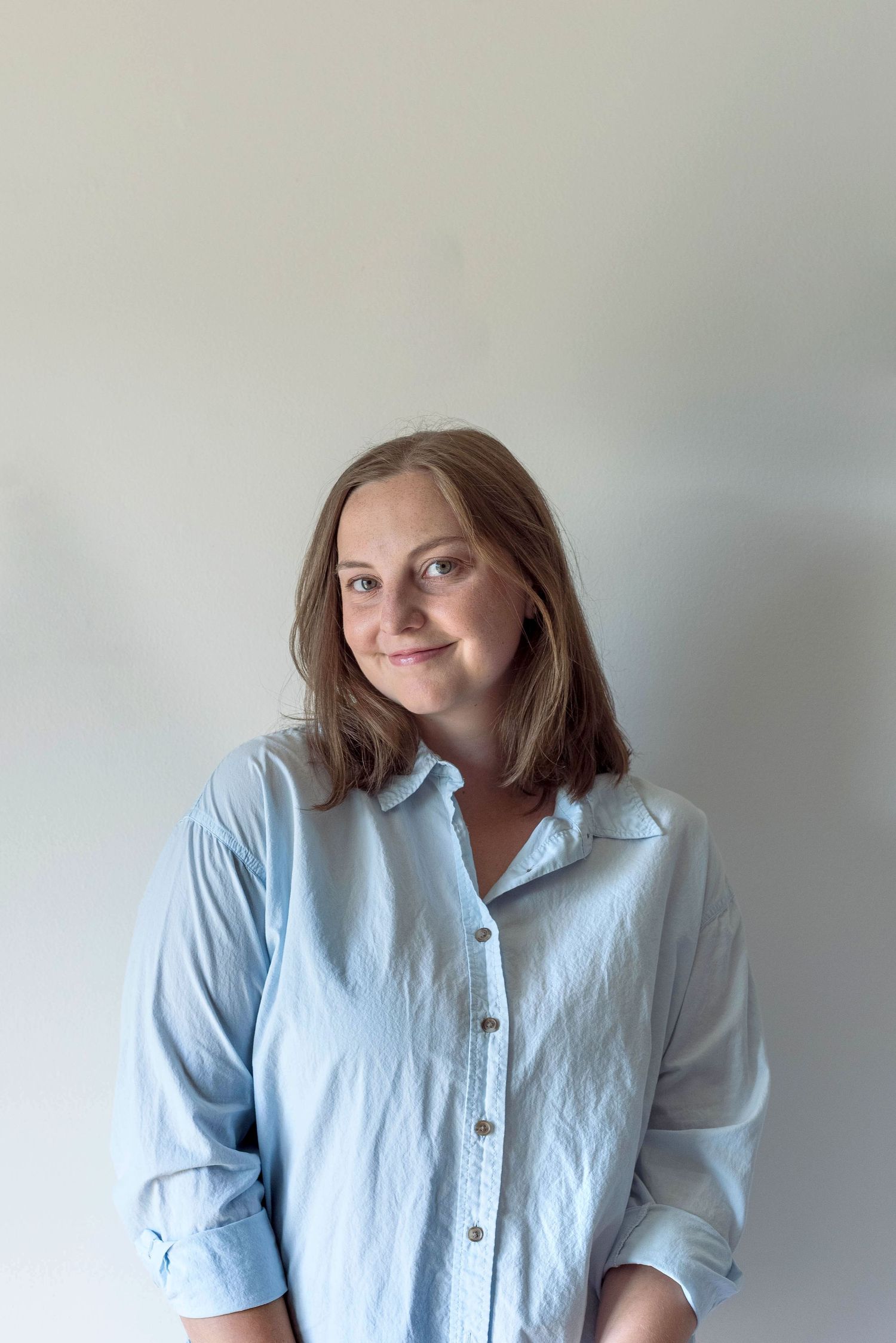 Young woman in light blue button-up shirt smiling softly against a white background.