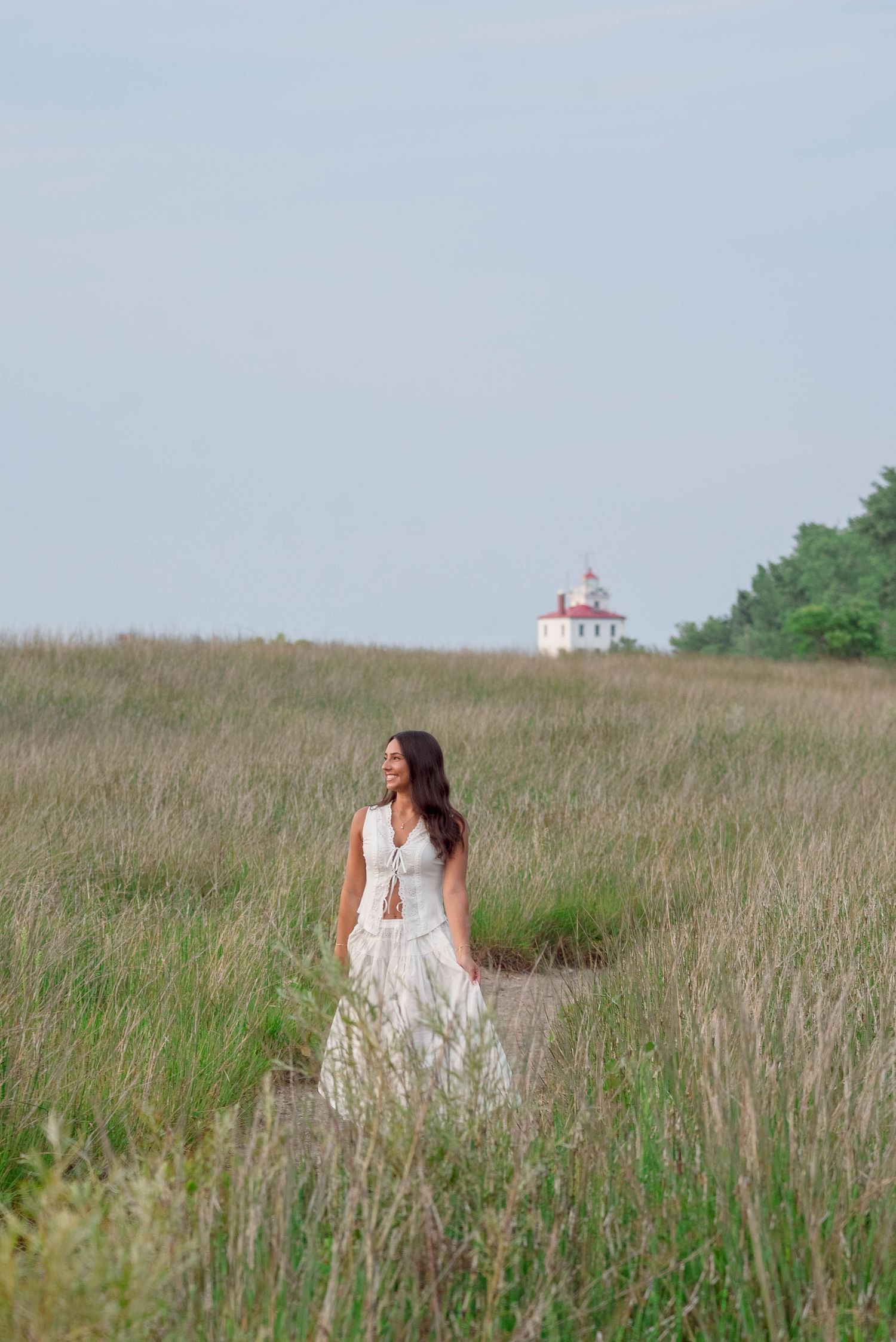 White dress portrait in tall grass with a lighthouse visible in the distance.