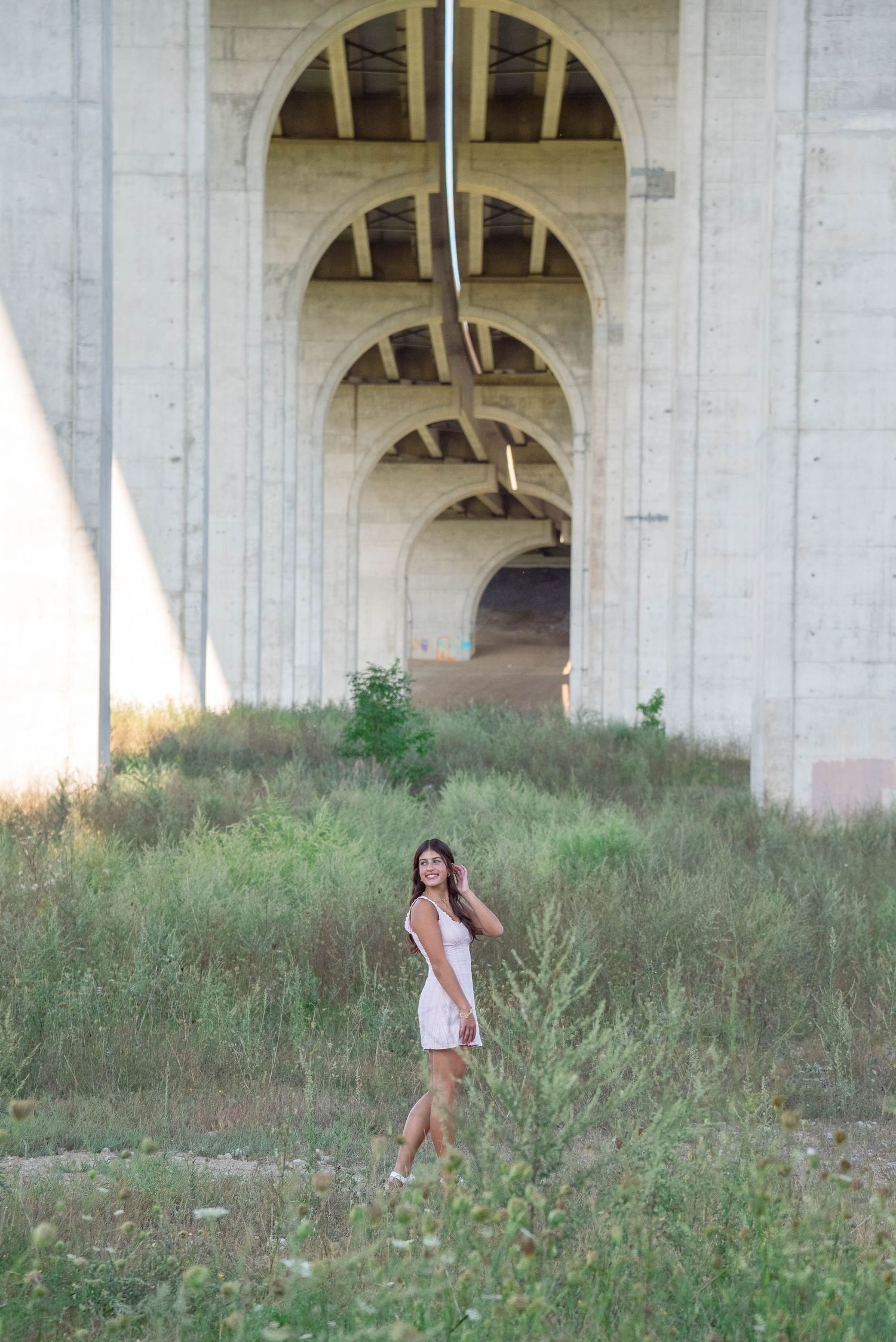 A white sundress under dramatic concrete bridge arches surrounded by wild grass.