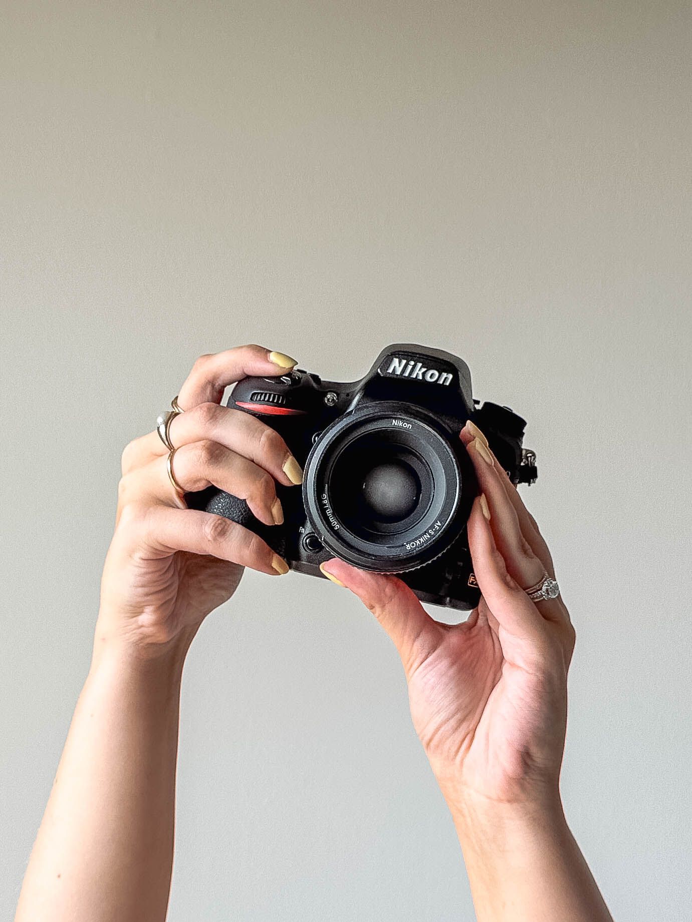 Hands holding a Nikon DSLR camera with a wide lens against a neutral gray background.
