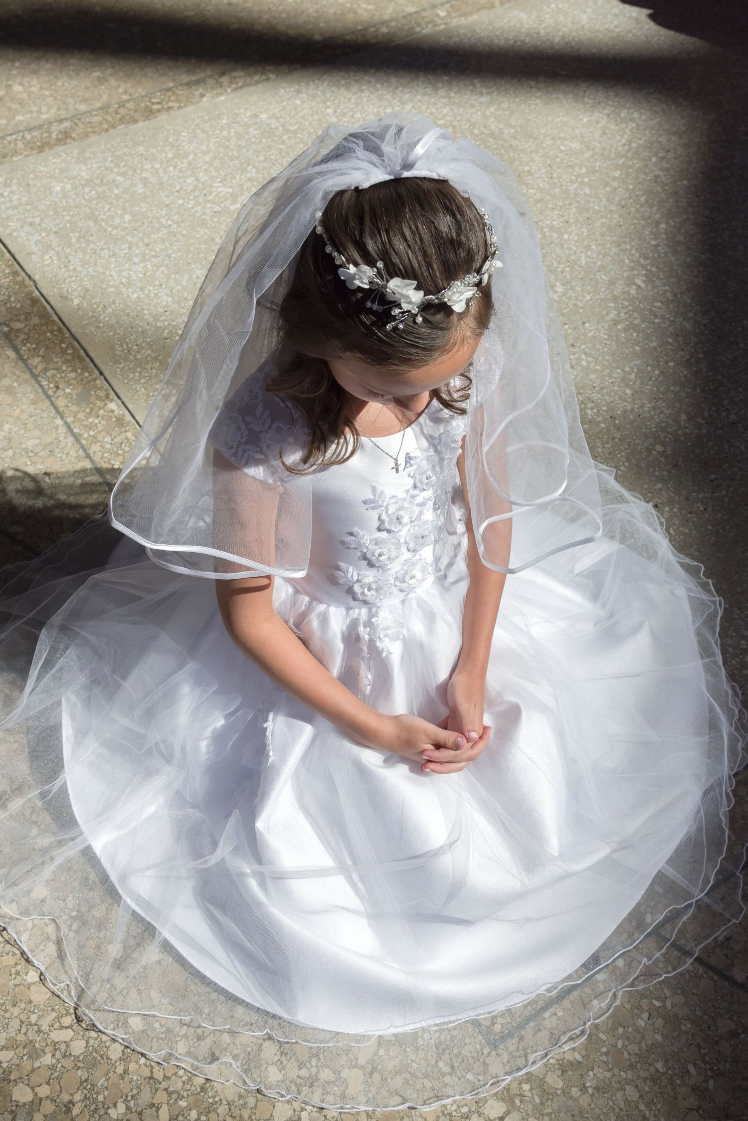 Young girl in white communion dress sitting gracefully on stone steps.