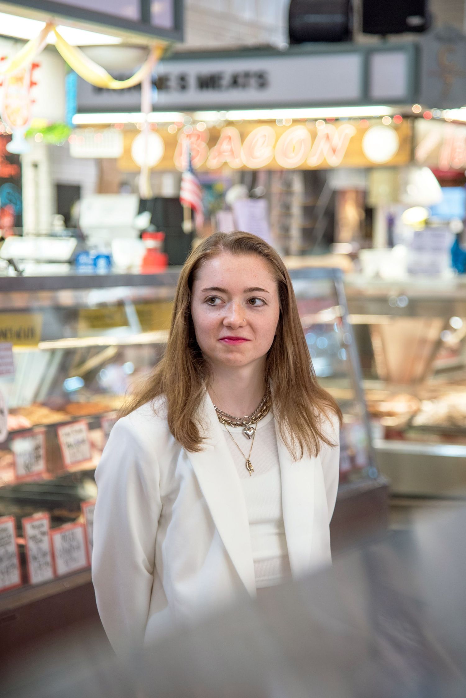 Person in white blazer photographed at market counter with neon signs in background.